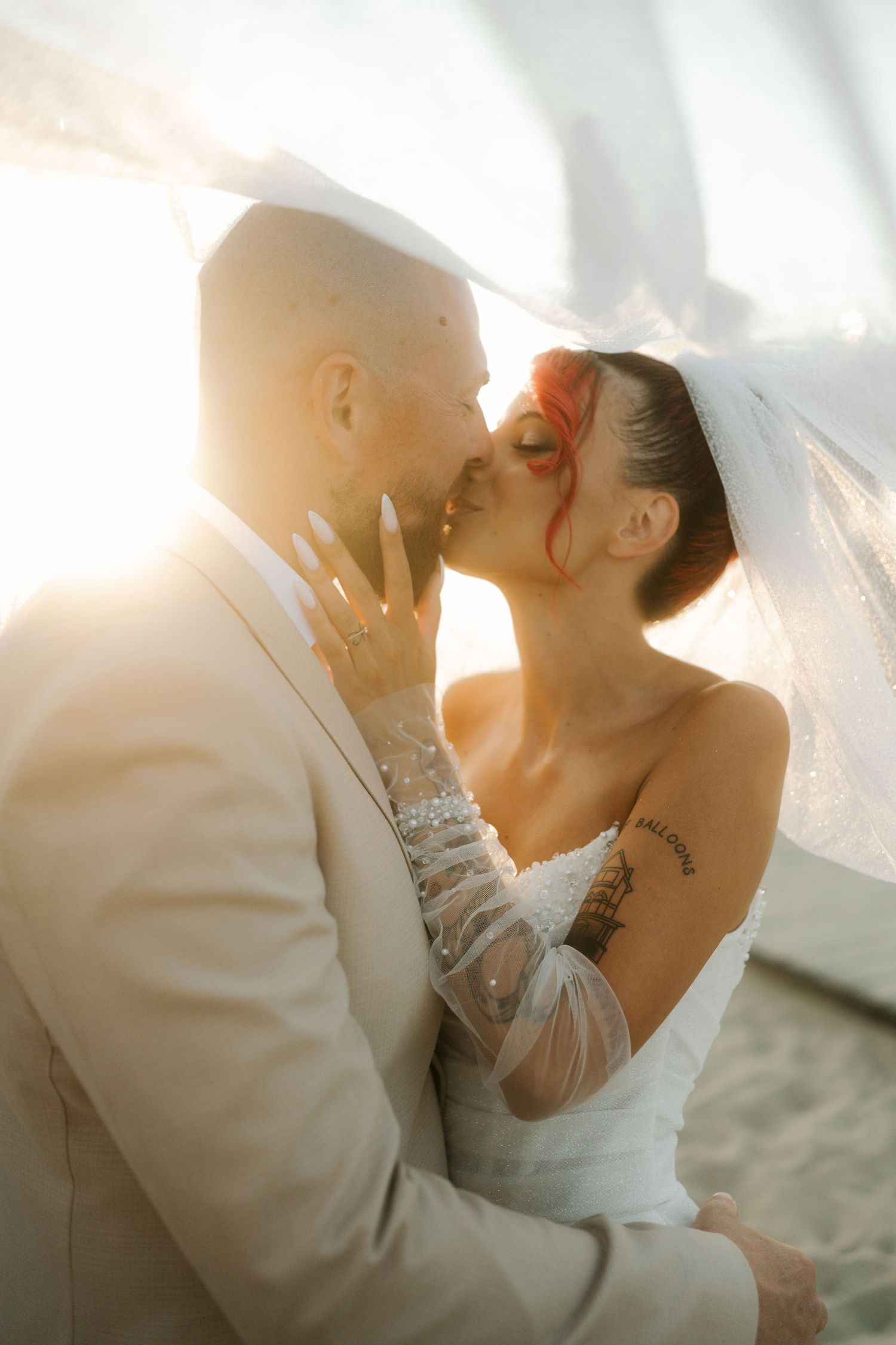 A romantic wedding couple shares a tender kiss under a soft glowing sunset light with a flowing veil in the breeze.