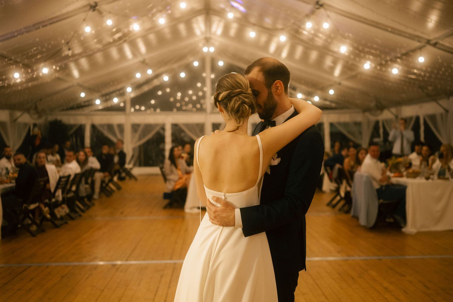 A couple shares their first dance in a beautifully lit wedding tent with string lights overhead and guests seated at tables.
