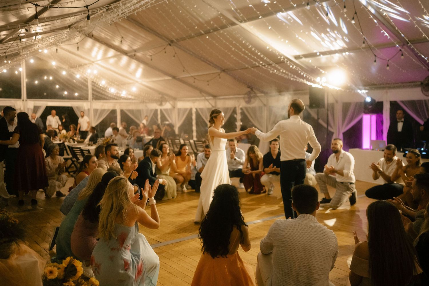 Wedding guests dance and celebrate under a glowing tent canopy at an evening reception with string lights overhead.