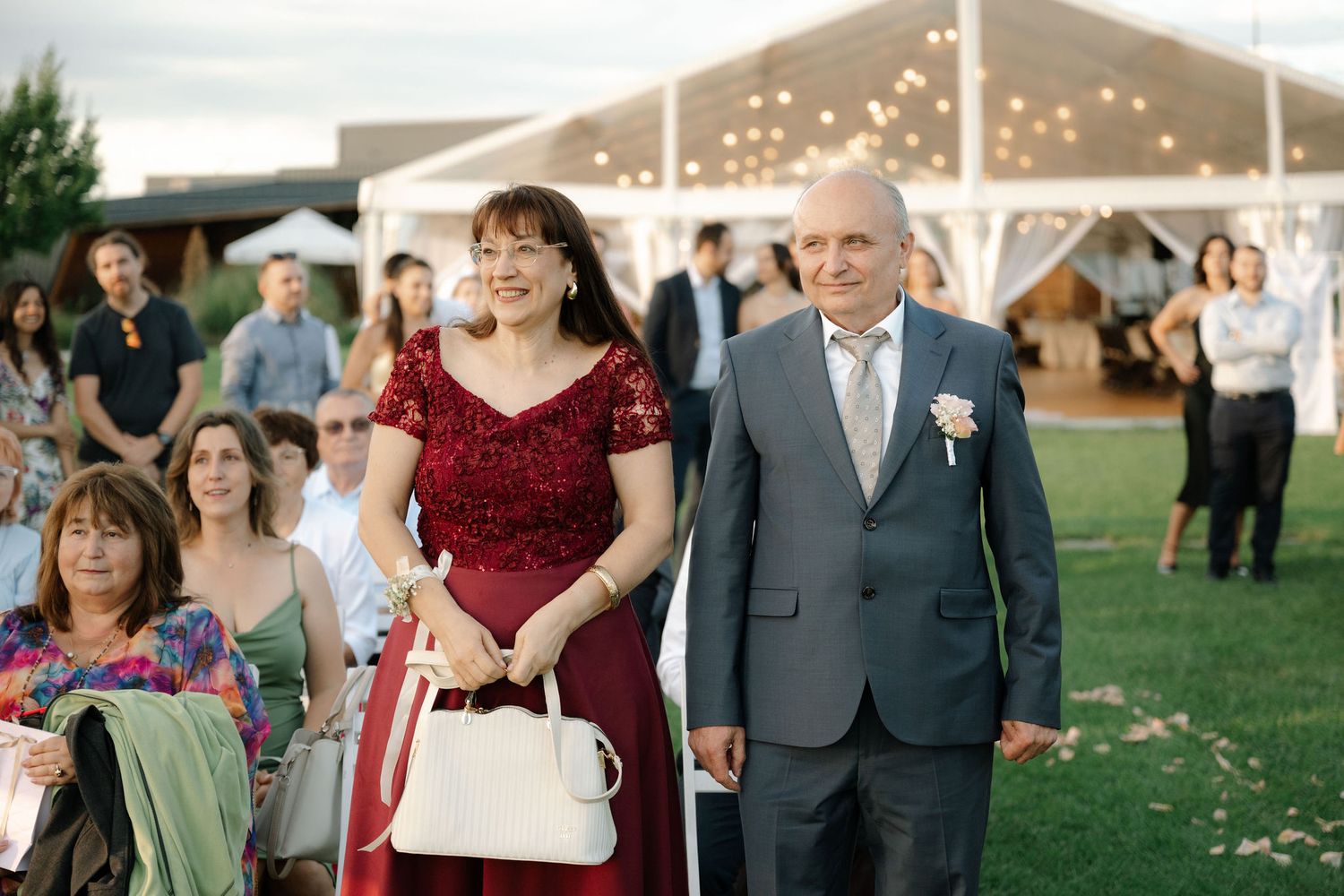 Guests at an outdoor wedding ceremony under a white tent with string lights during daytime.