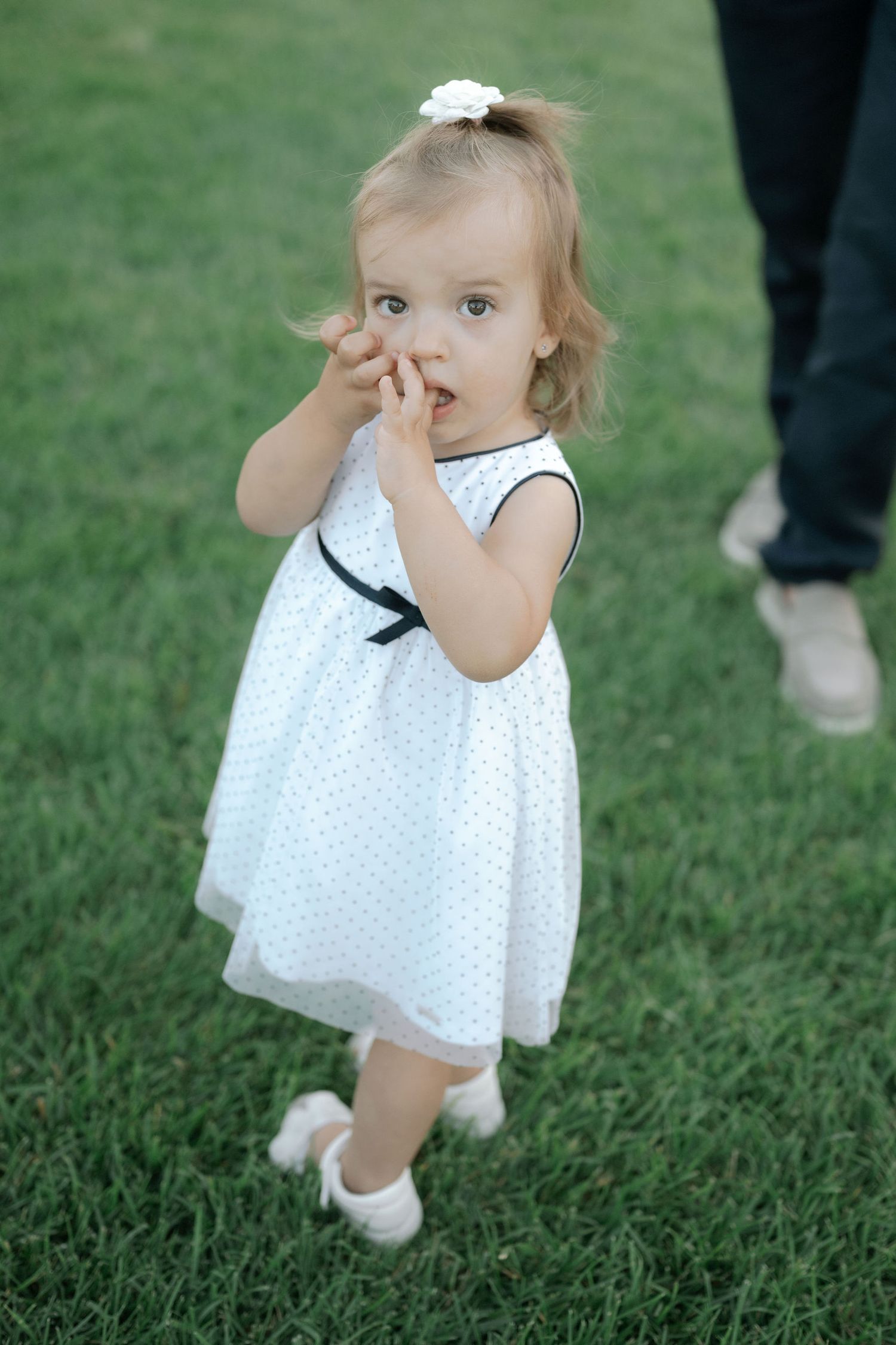 A small toddler in a white polka dot dress walks on grass while covering their face with their hand.