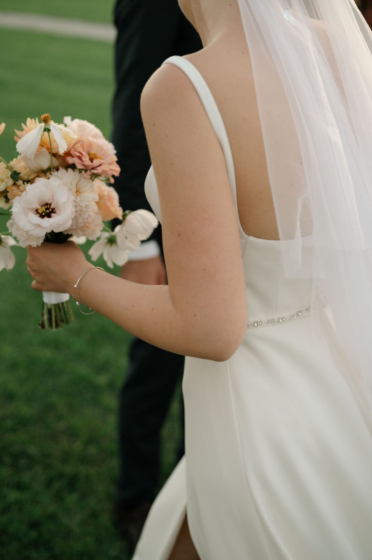 Bride in white wedding dress holds a delicate peach and white floral bouquet against a green outdoor backdrop.