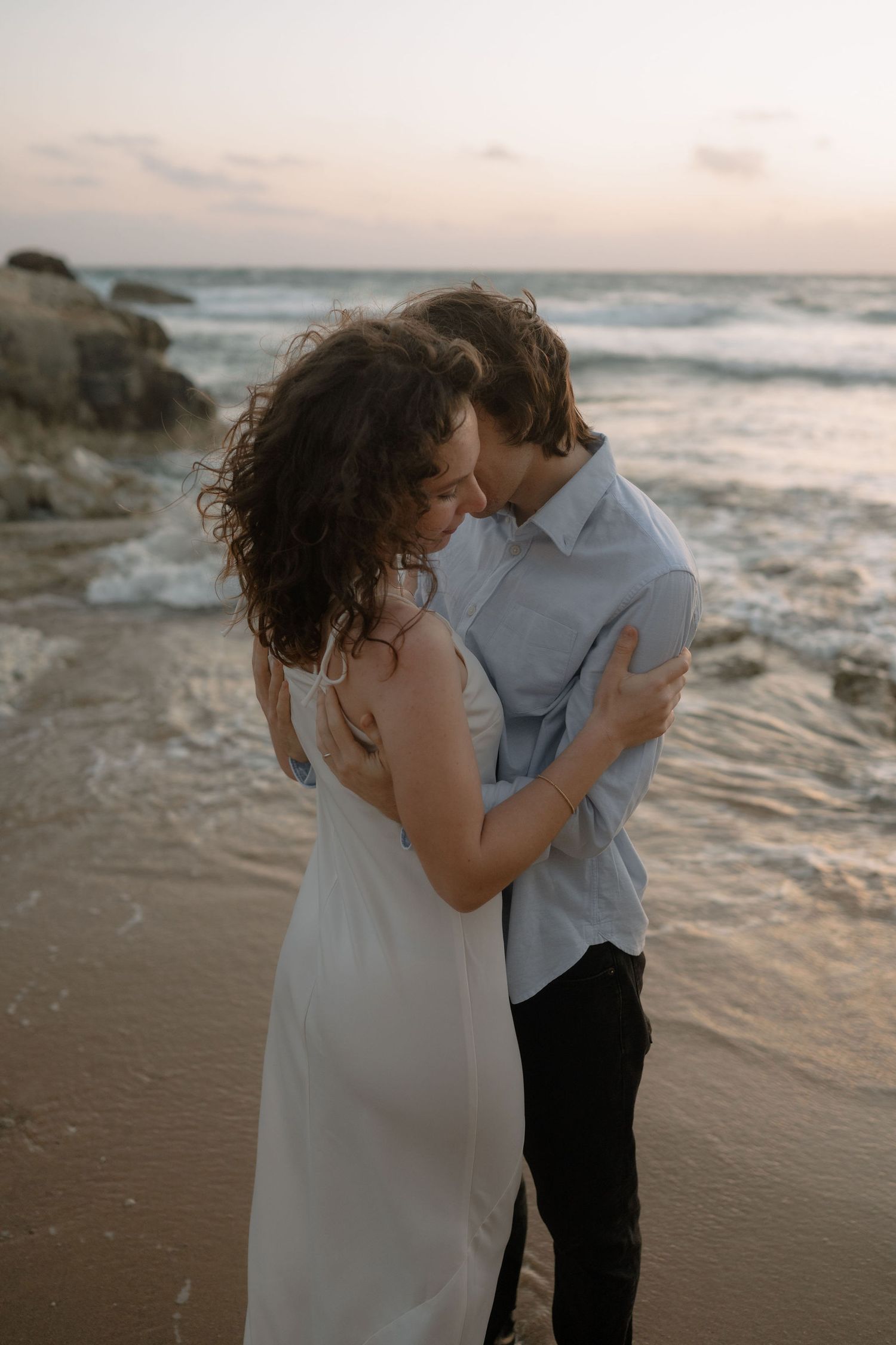 Couple embracing romantically on a sandy beach at sunset with ocean waves in the background.