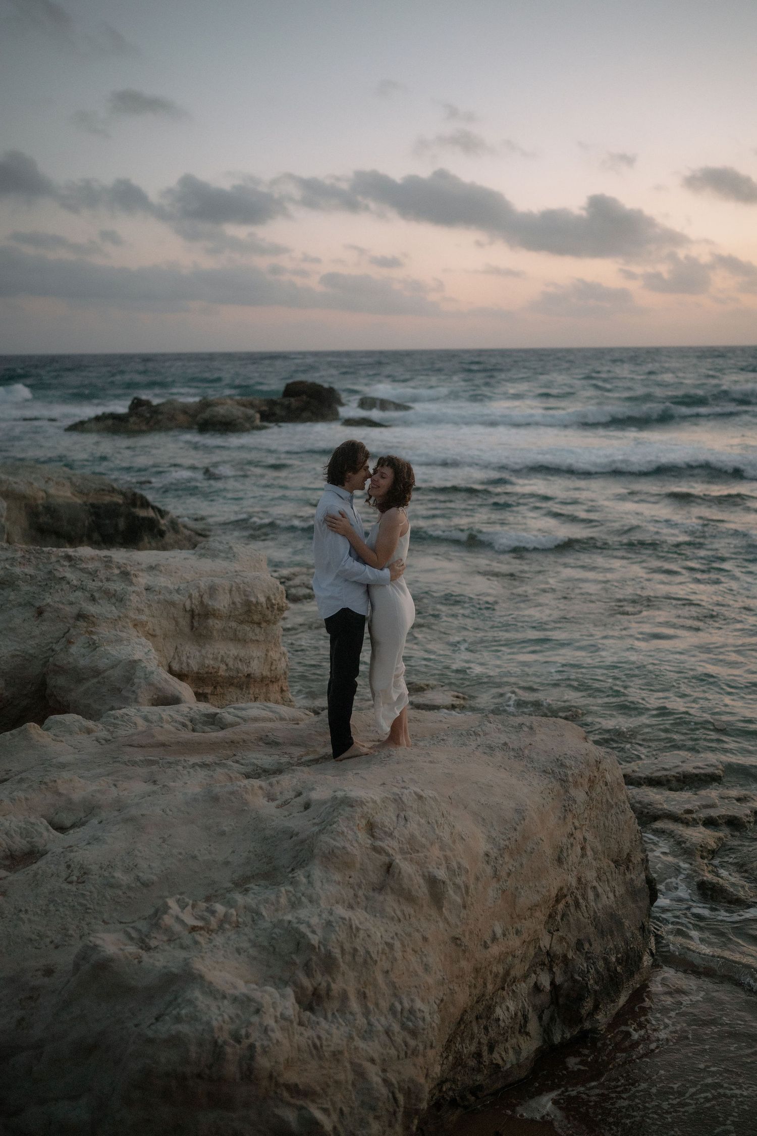 Couple embracing and kissing on rocky shoreline at sunset with ocean waves in background.