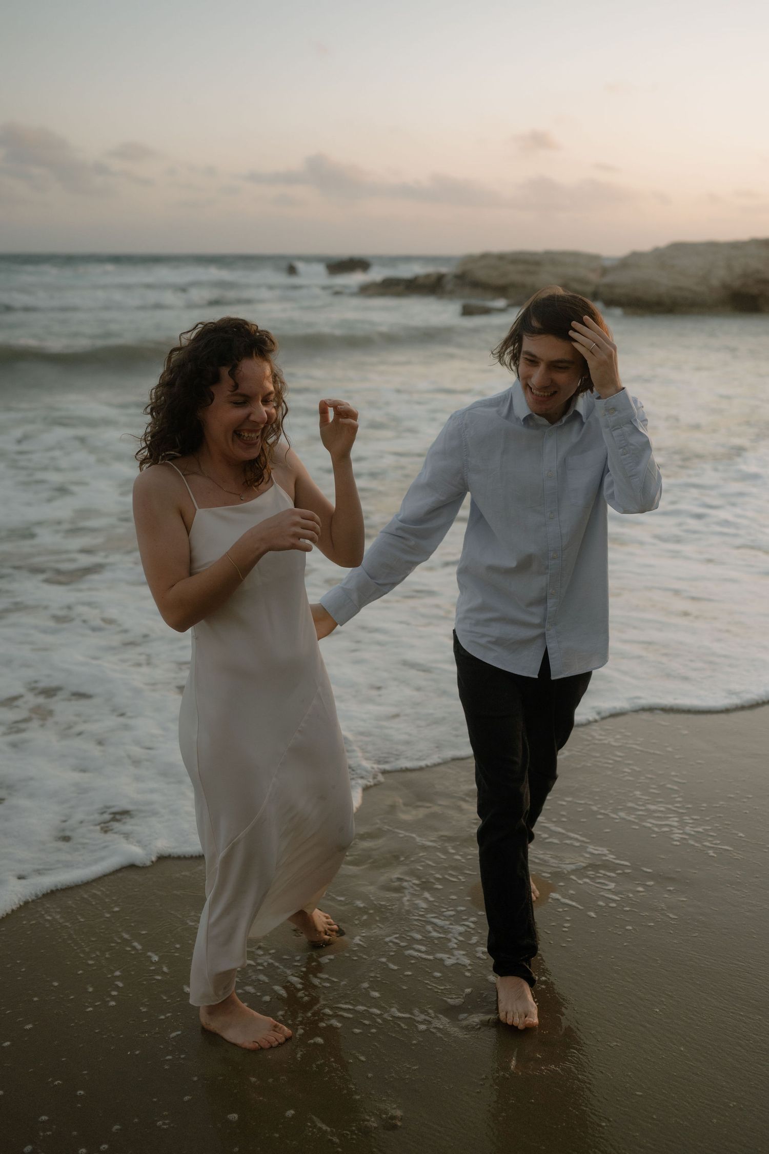Couple laughing and holding hands while walking barefoot on a sandy beach at sunset.