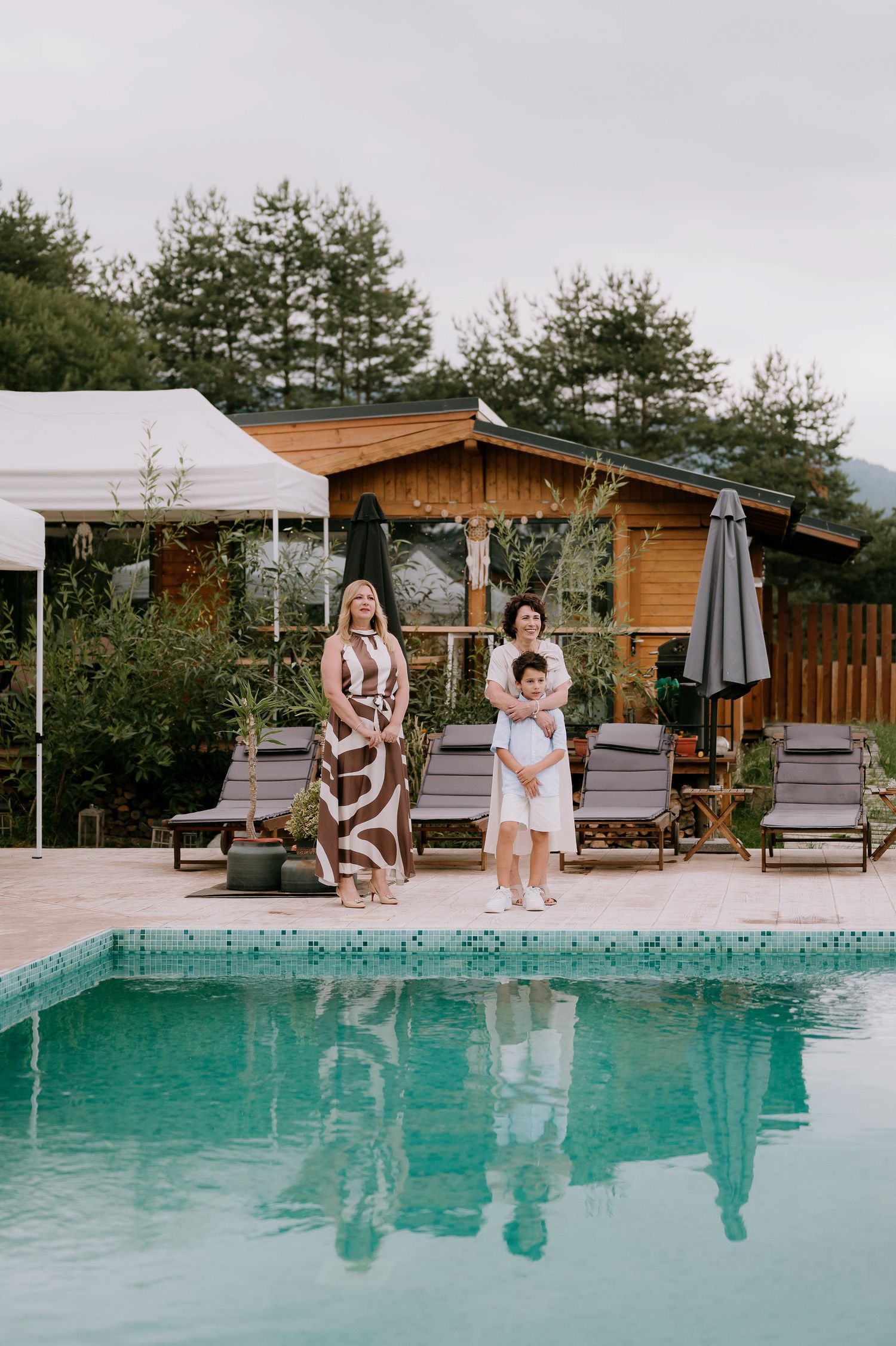 Guests relaxing by a turquoise pool surrounded by wooden lounge chairs and pine trees.
