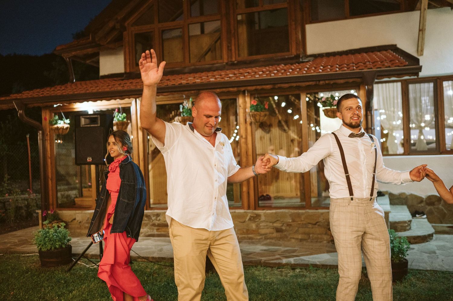 Wedding party in light colored attire dances outdoors near rustic barn.