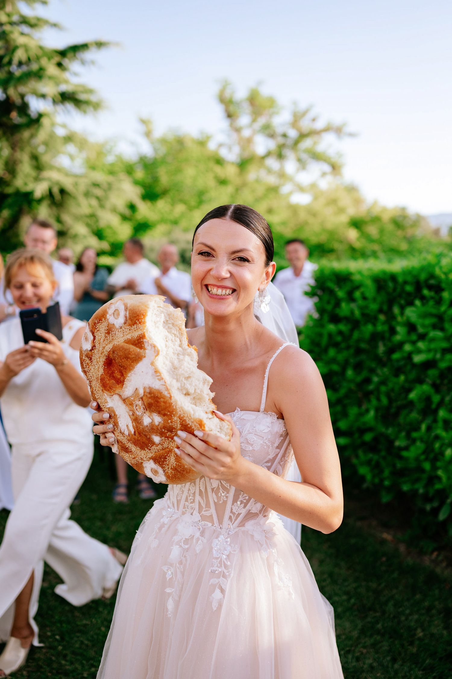Person in a wedding dress joyfully displays a traditional round loaf of bread at an outdoor reception.