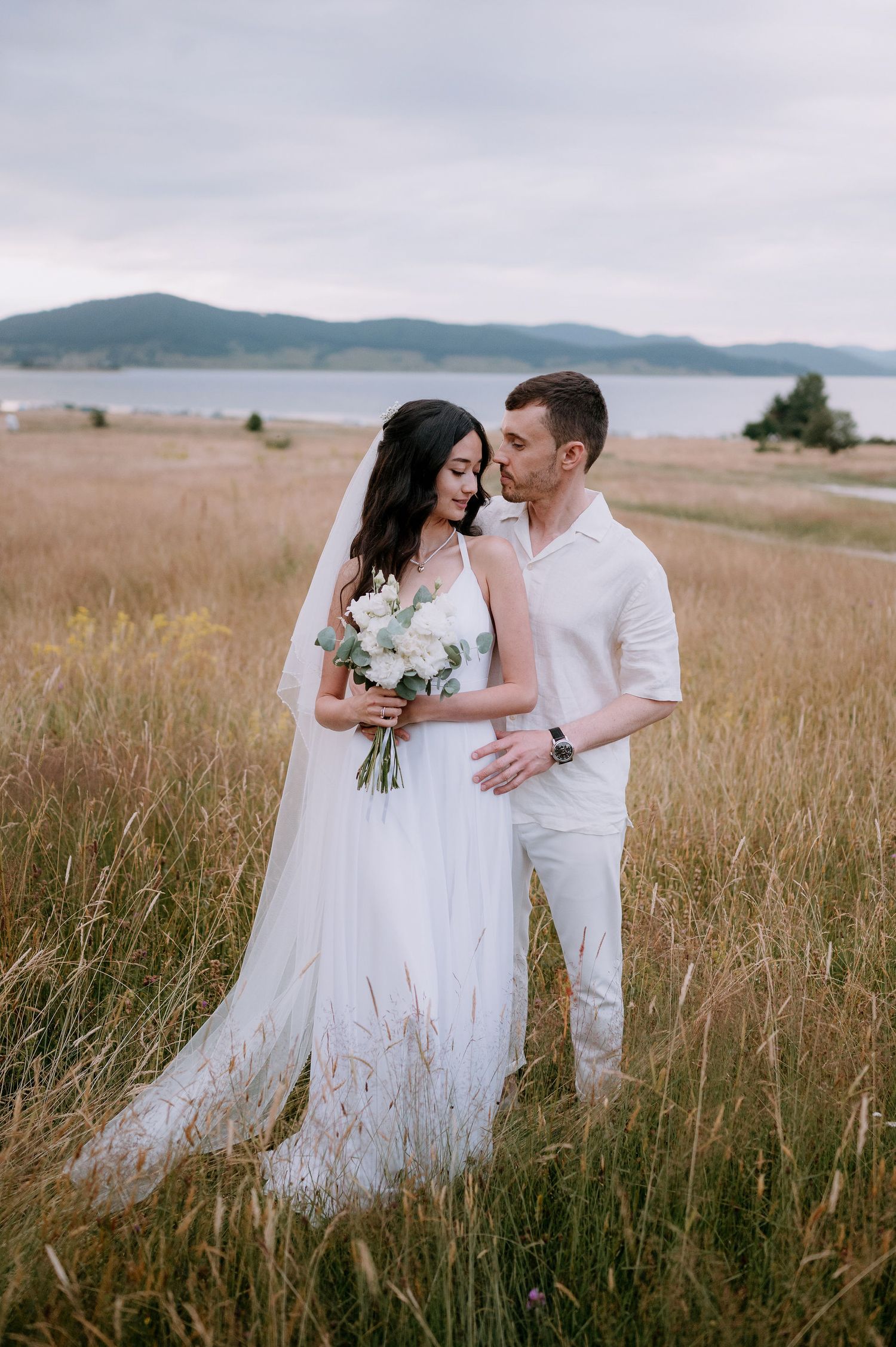 Bride and groom embrace in flowing white attire against mountainous landscape.