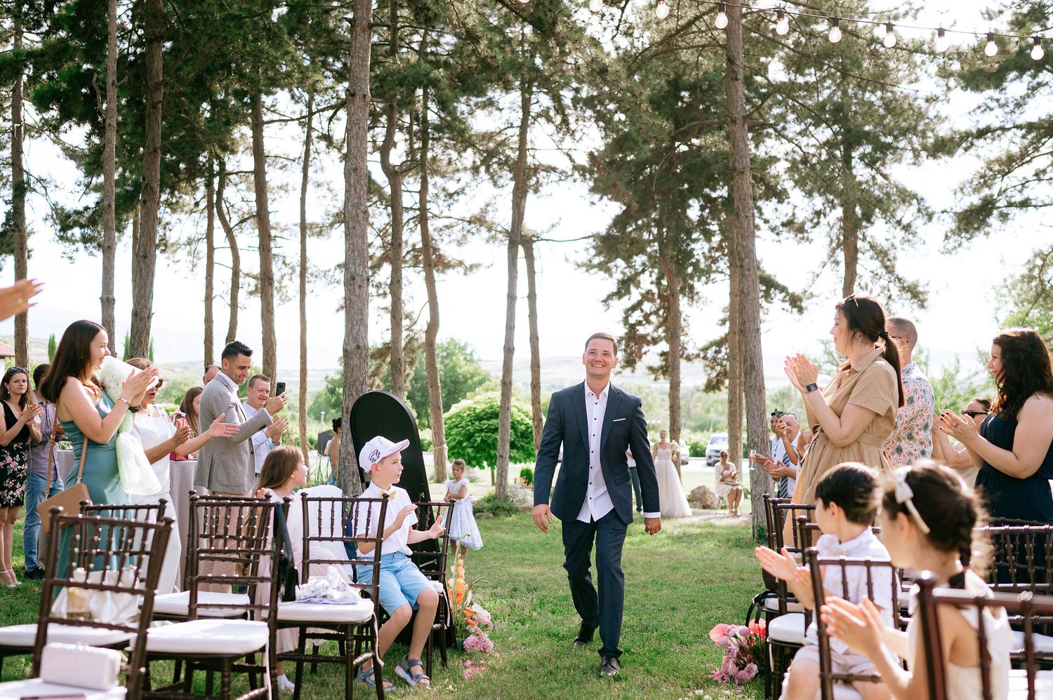 Outdoor wedding ceremony with guests seated on wooden chairs among tall pine trees.