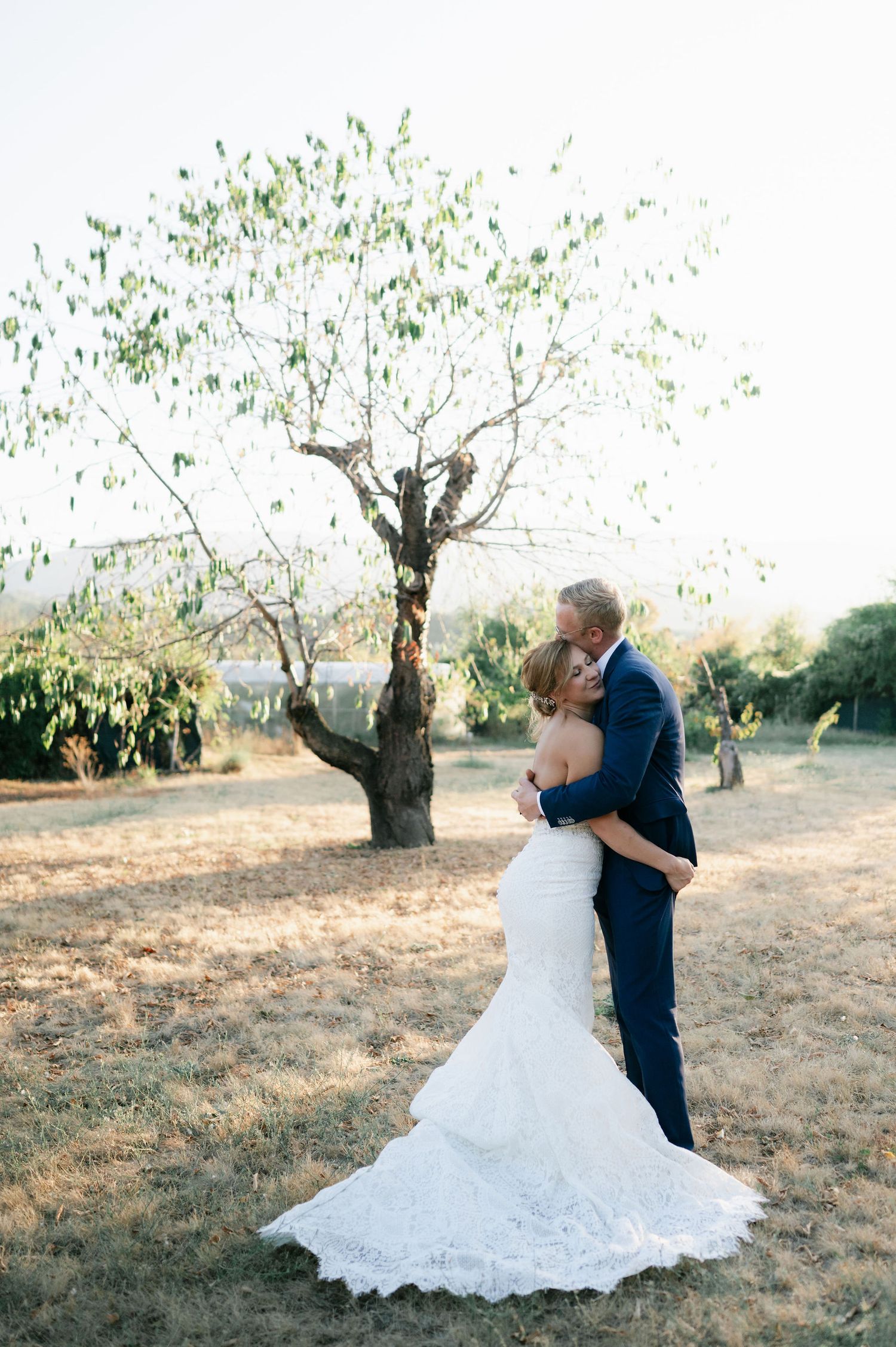A couple dances together in an olive grove during golden hour, her white wedding dress flowing in the sunlight.