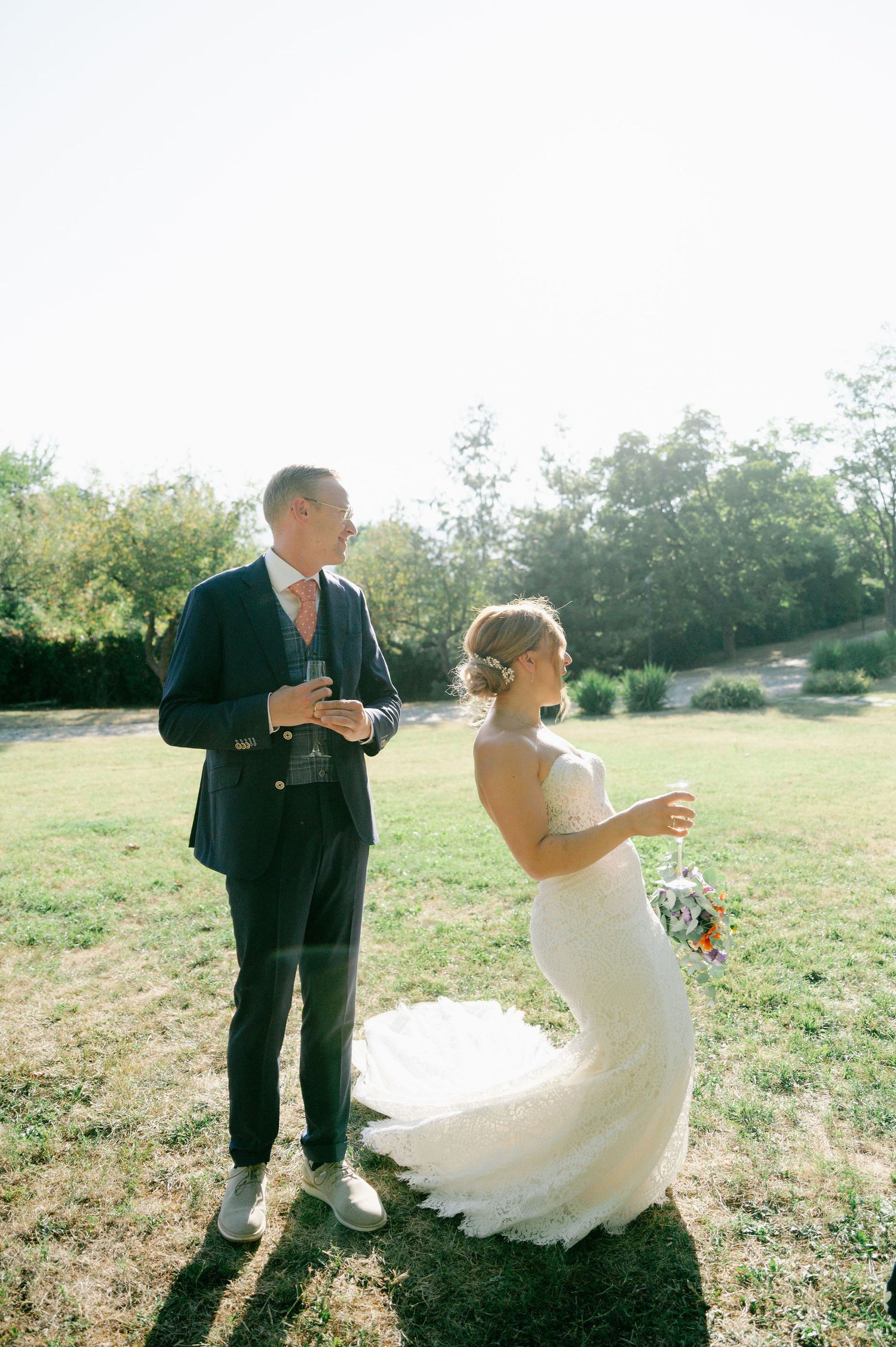 A couple in wedding attire spins playfully in a sunlit meadow.