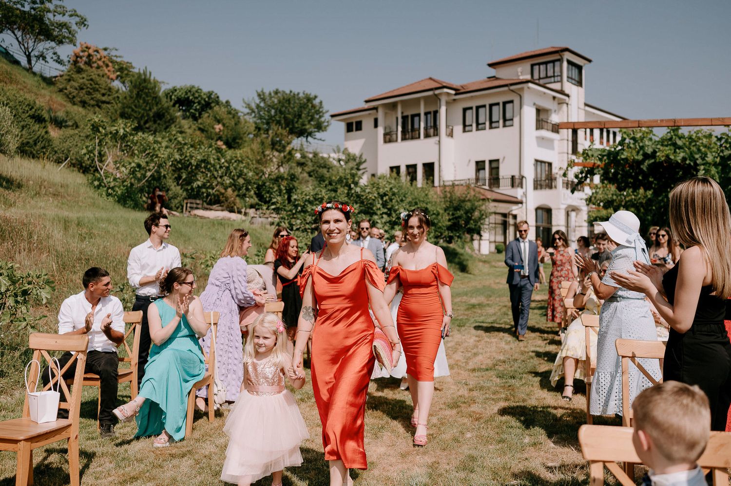 Wedding guests seated in wooden chairs on a hillside with a white building in the background.