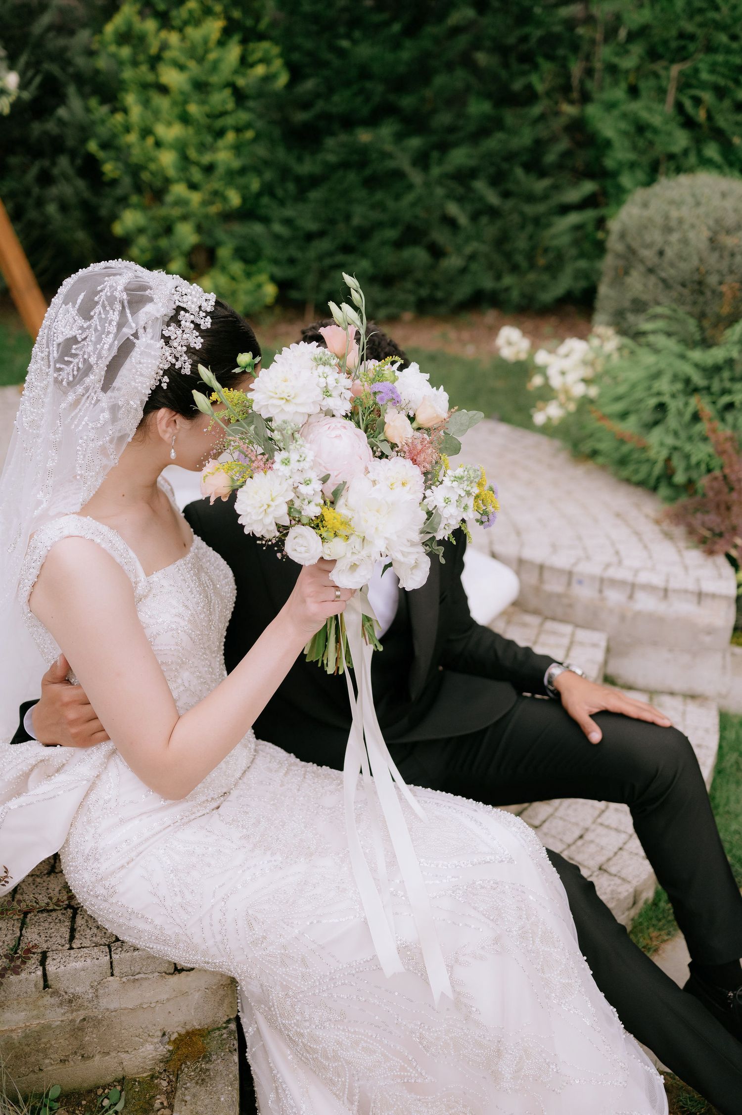 Romantic wedding couple sitting on stone wall surrounded by lush garden scenery.