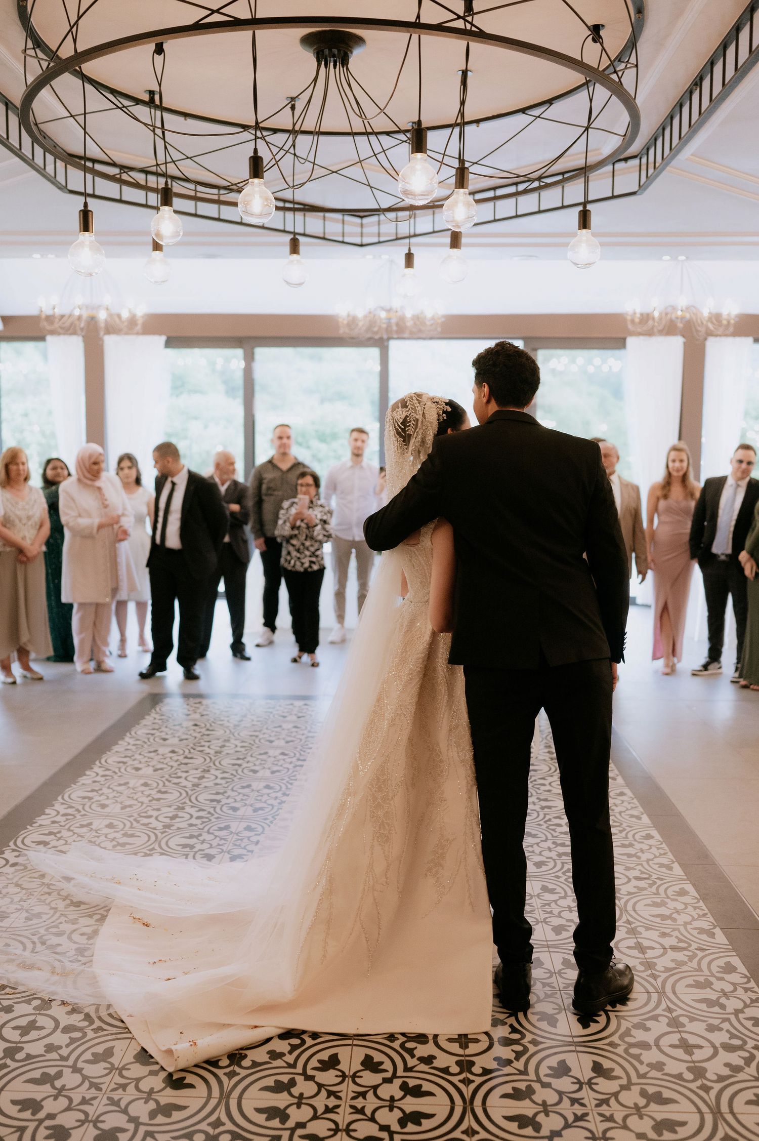 A wedding couple dances together on an ornate patterned floor in an elegant reception venue with hanging lights.