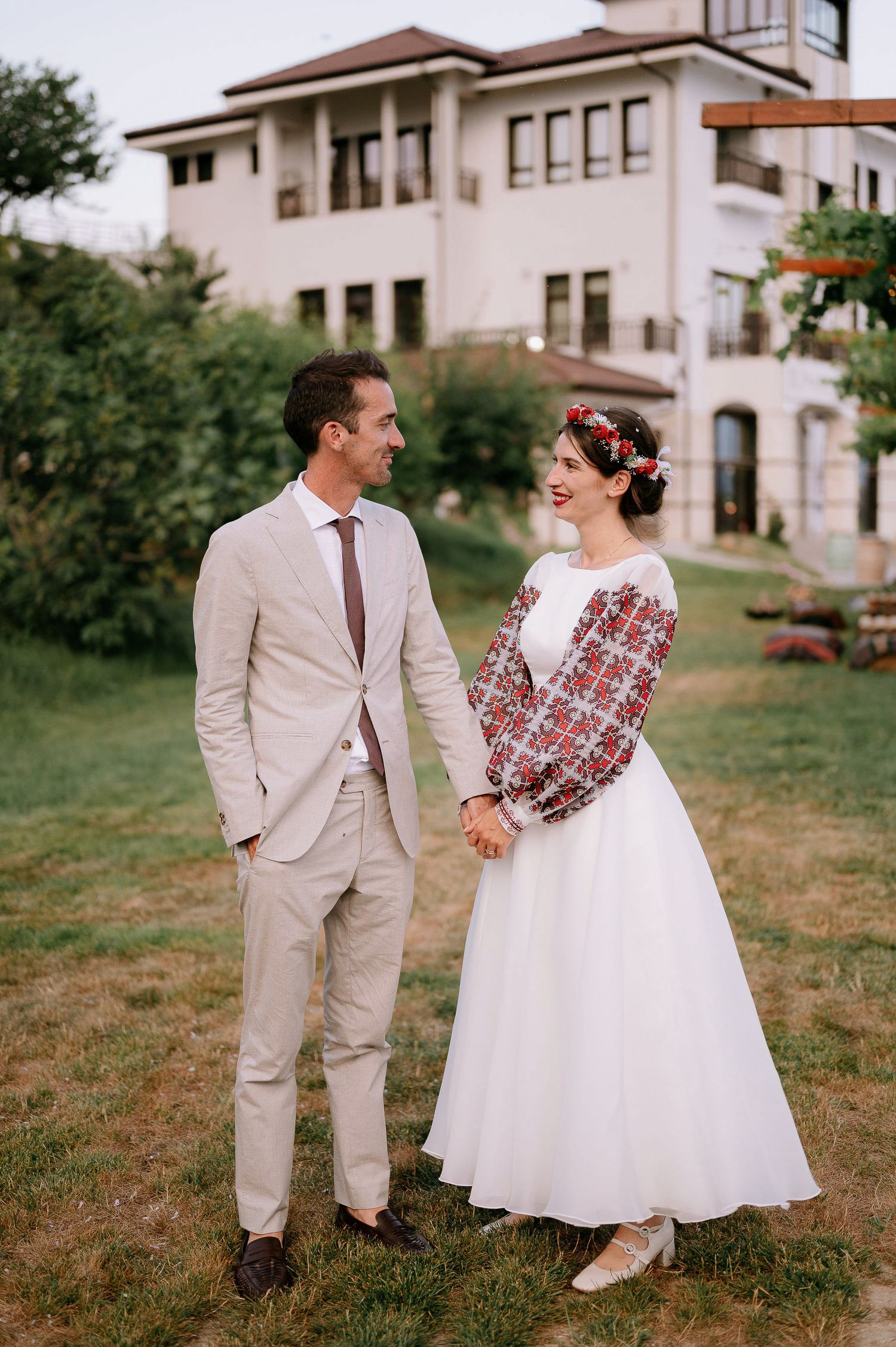 A couple in wedding attire walking and smiling on a grassy hill near a Mediterranean-style building.
