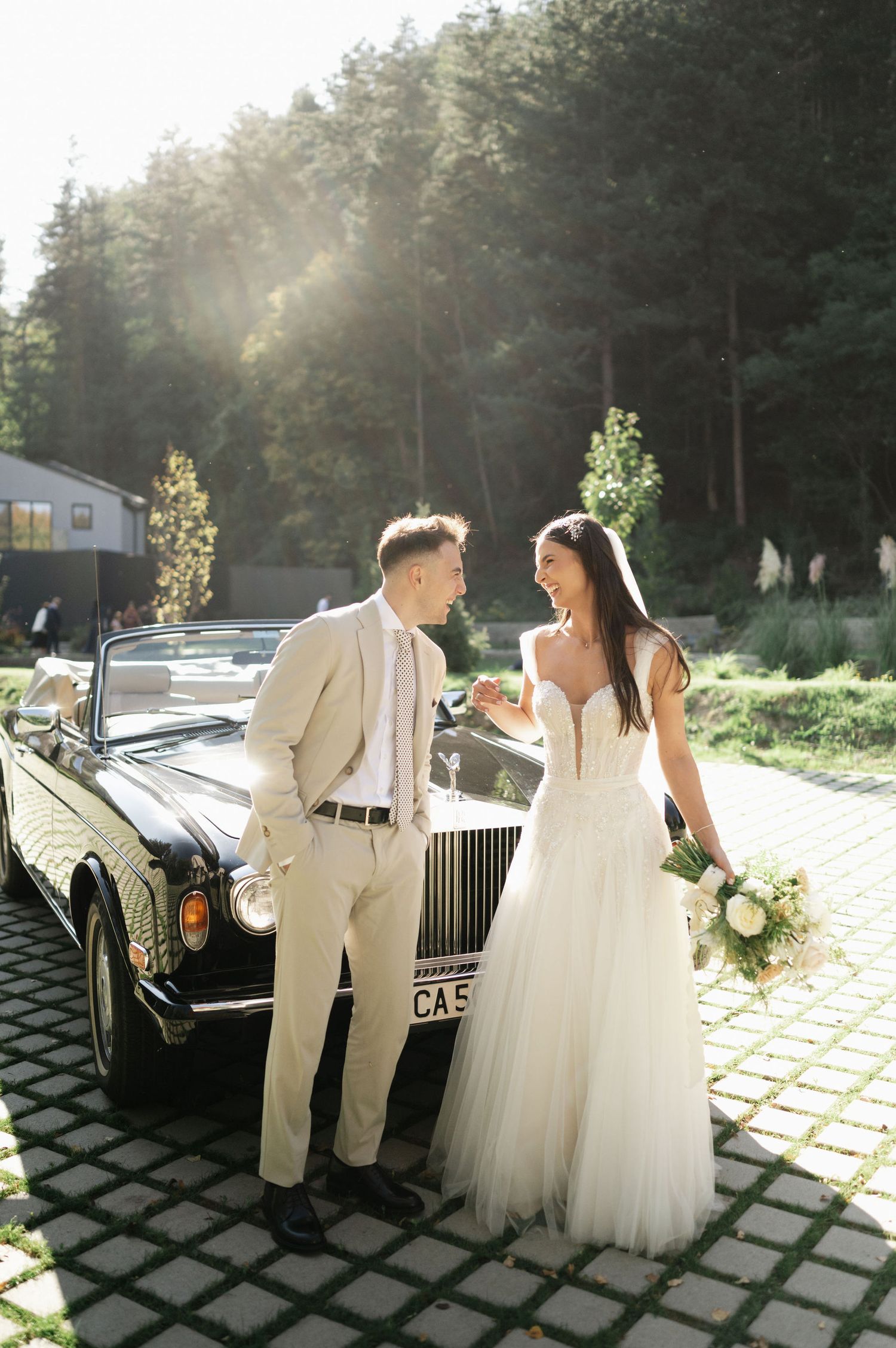 Romantic wedding moment captured beside luxury vintage car with sunlit forest background.
