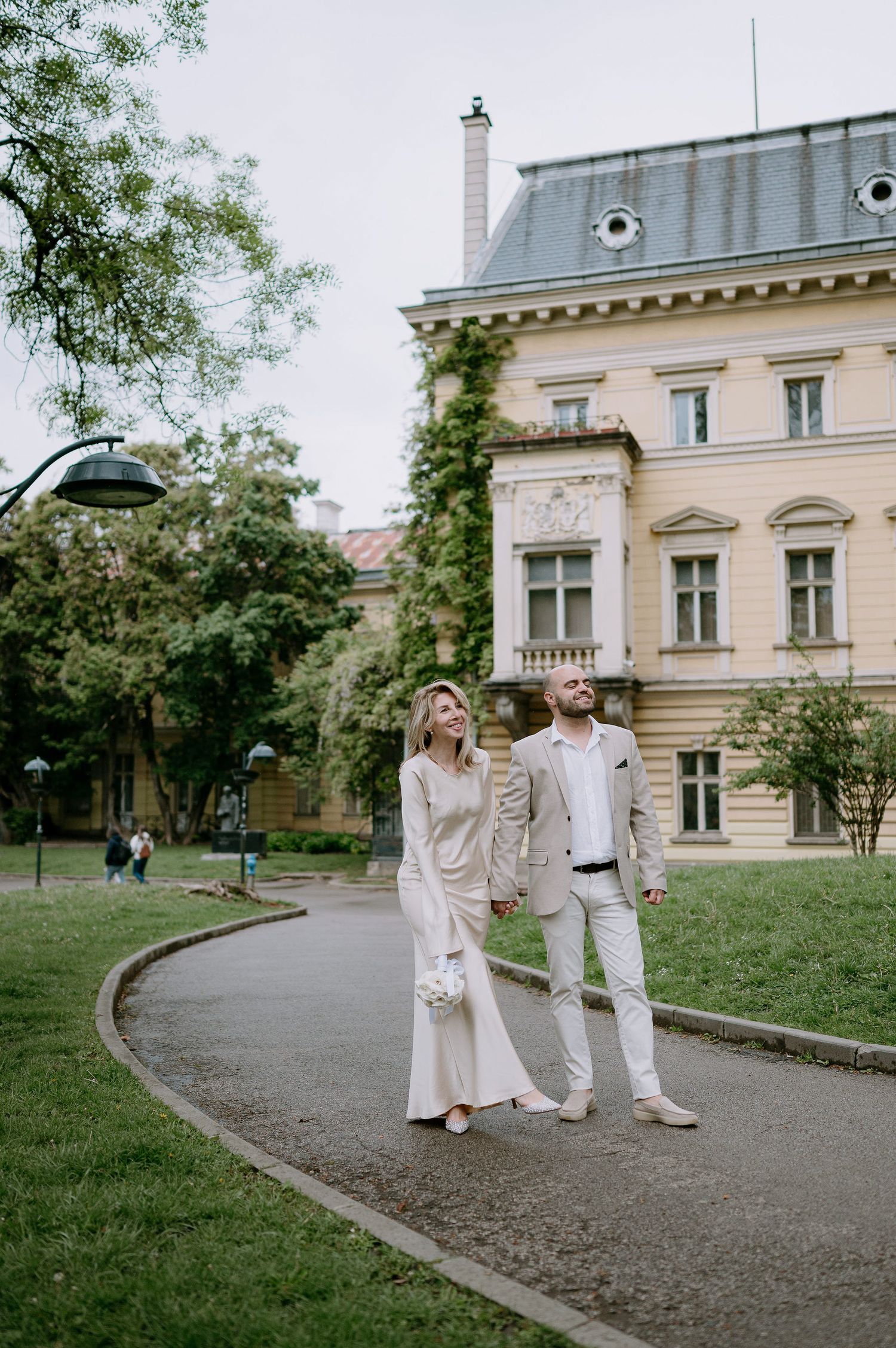 A couple in matching white outfits walk along a curved path near a historic building.