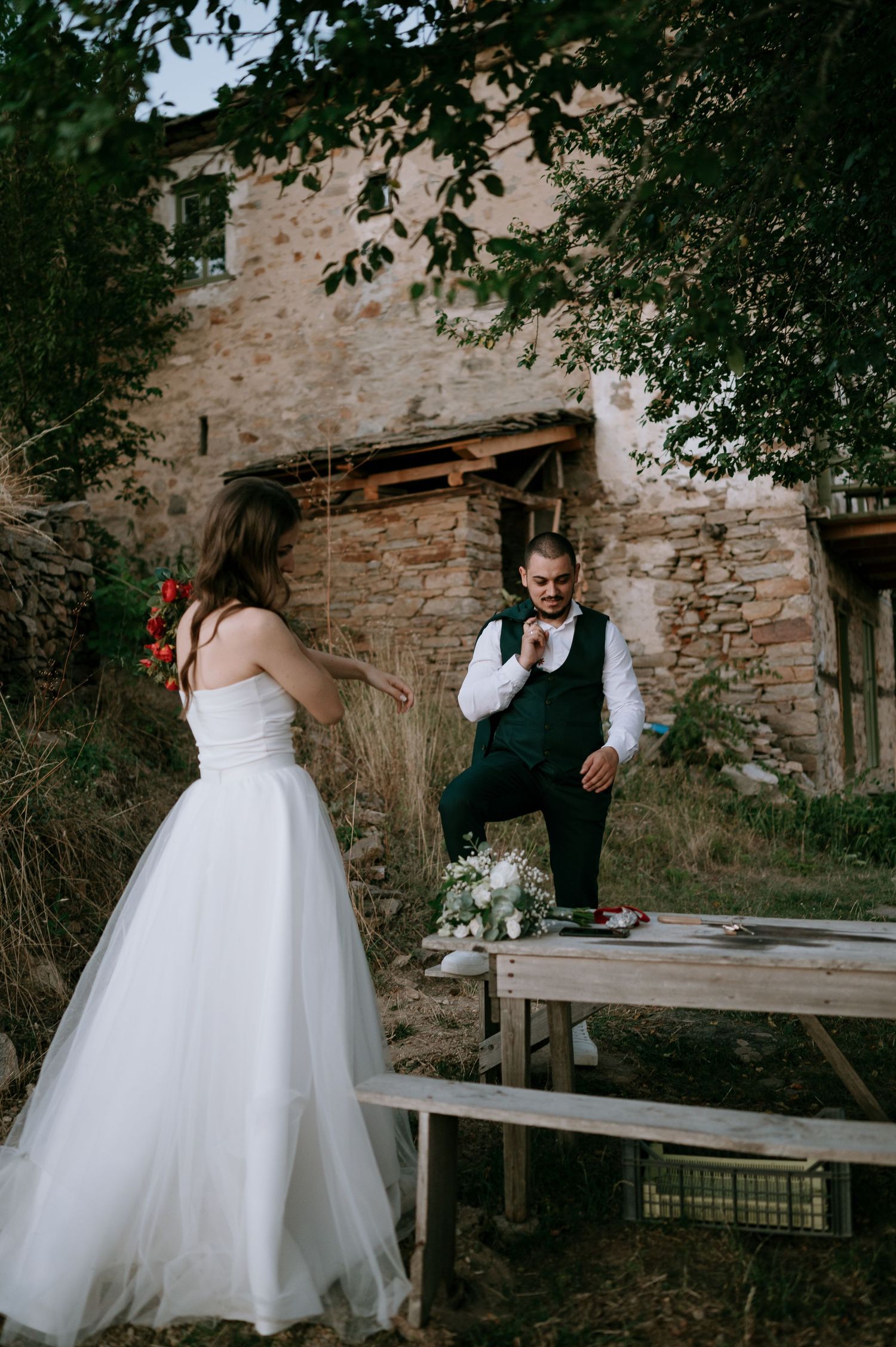 Wedding couple interacting playfully on wooden benches near rustic stone building.