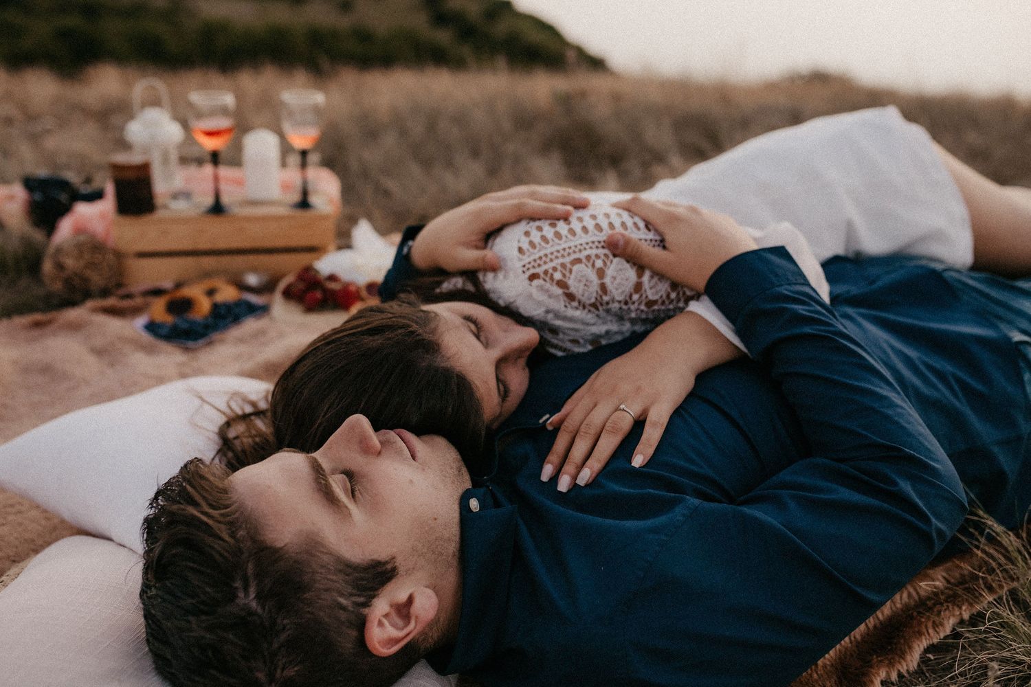 Couple lying on blanket during mountain picnic with wine glasses nearby.