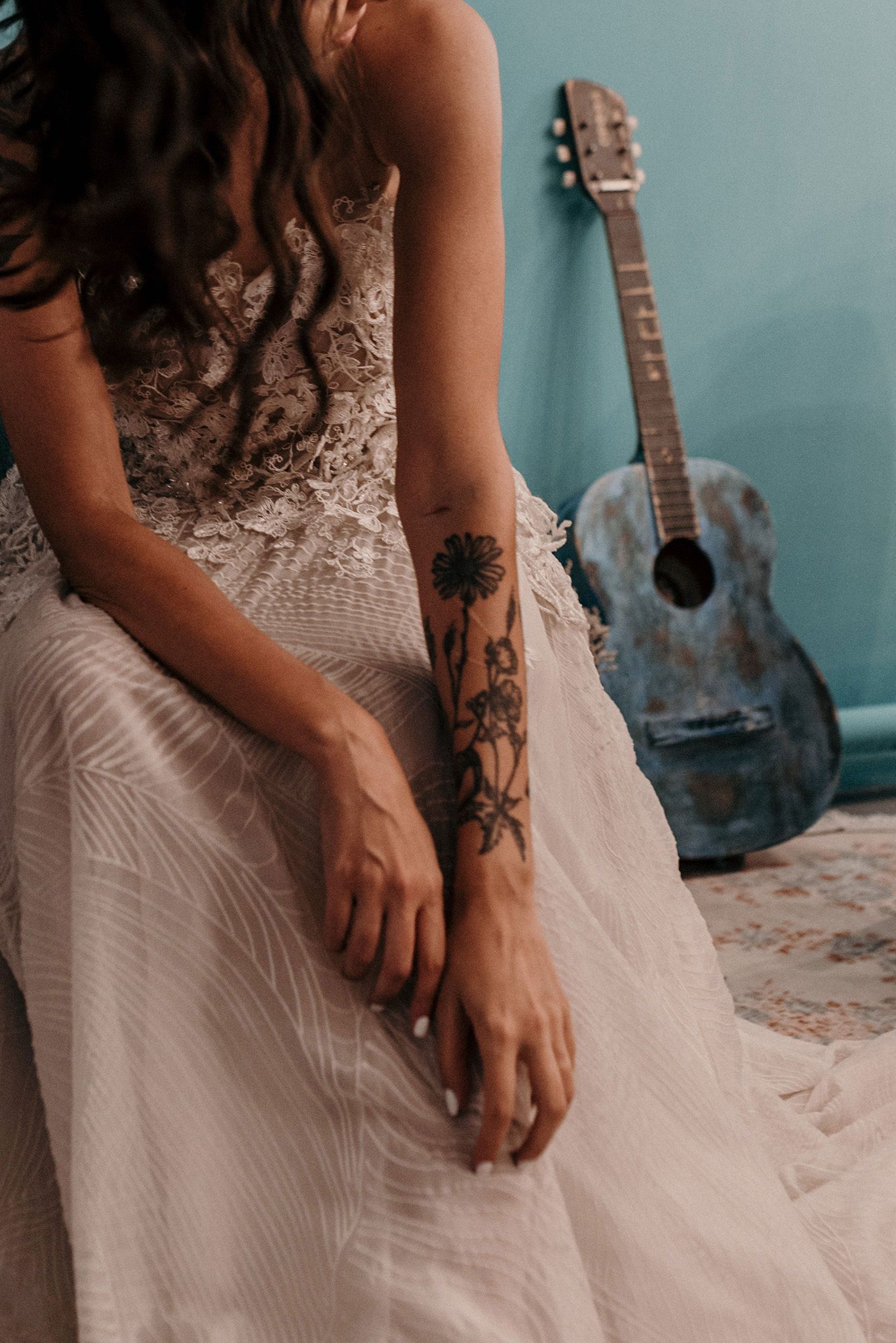 Close-up of floral tattoos on arm with white lace wedding dress and acoustic guitar in background.