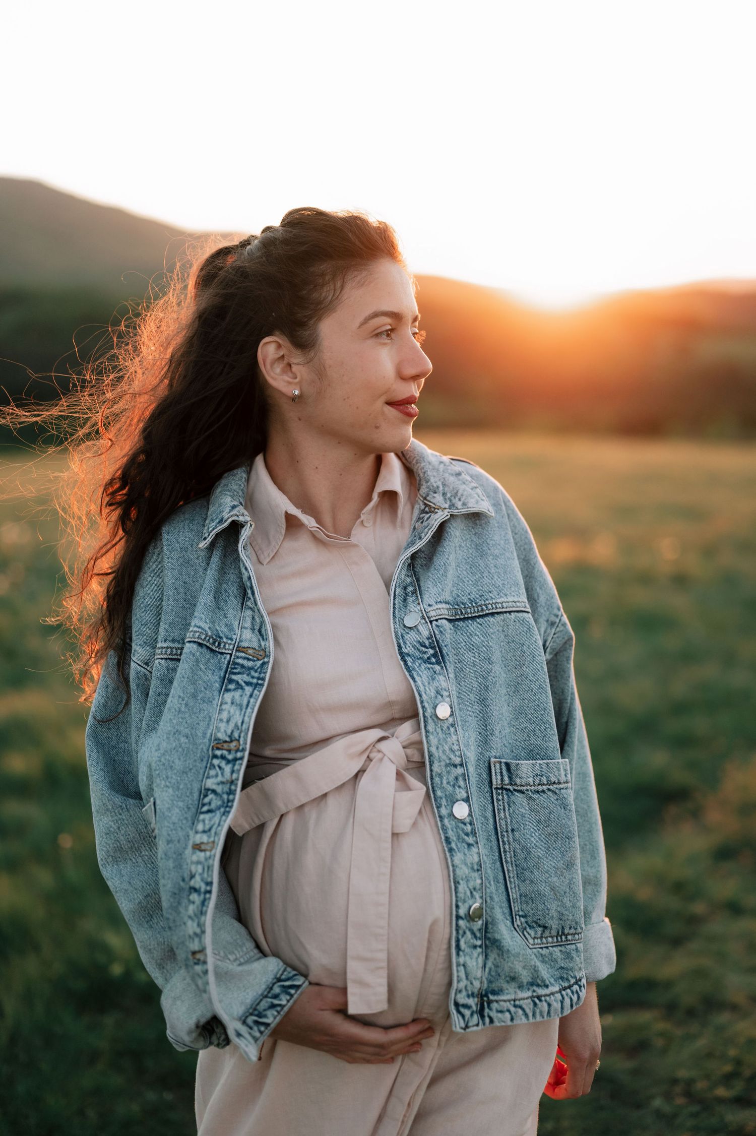 Woman in denim jacket and flowing dress stands against golden sunset backdrop.