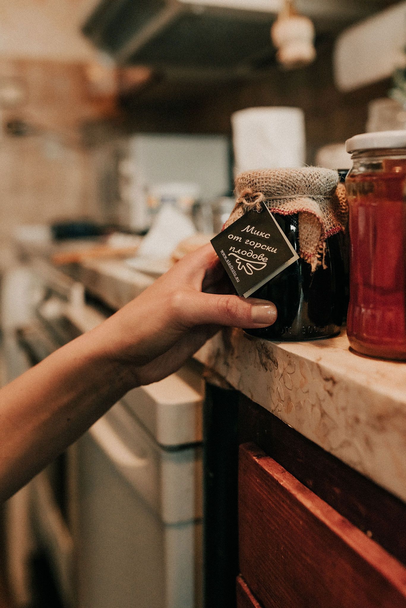 A hand reaches for a rustic jar with burlap top on a wooden shelf, with red preserves visible in background.