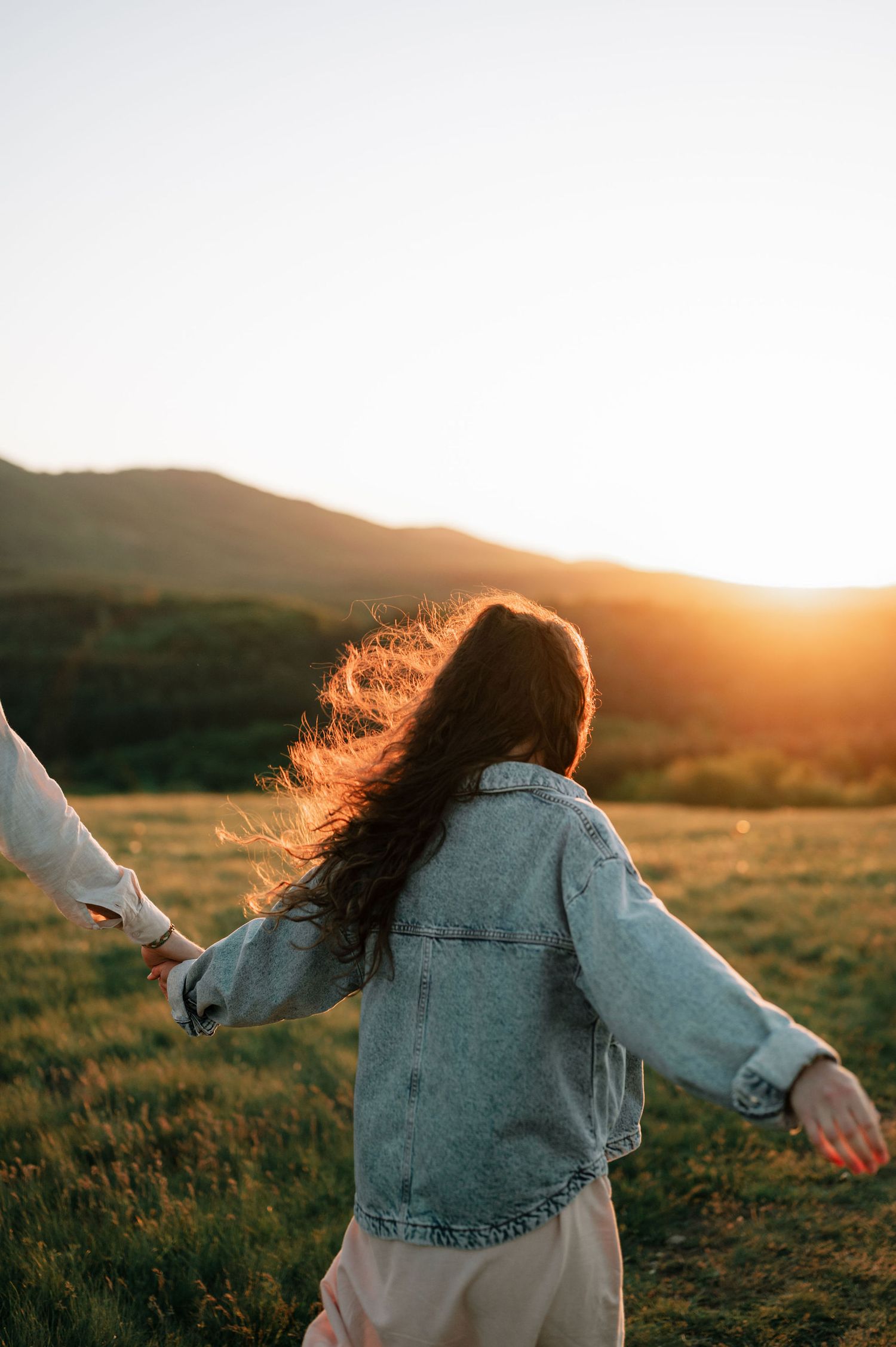 Person in denim jacket twirls with arms outstretched in a field during golden hour sunset.