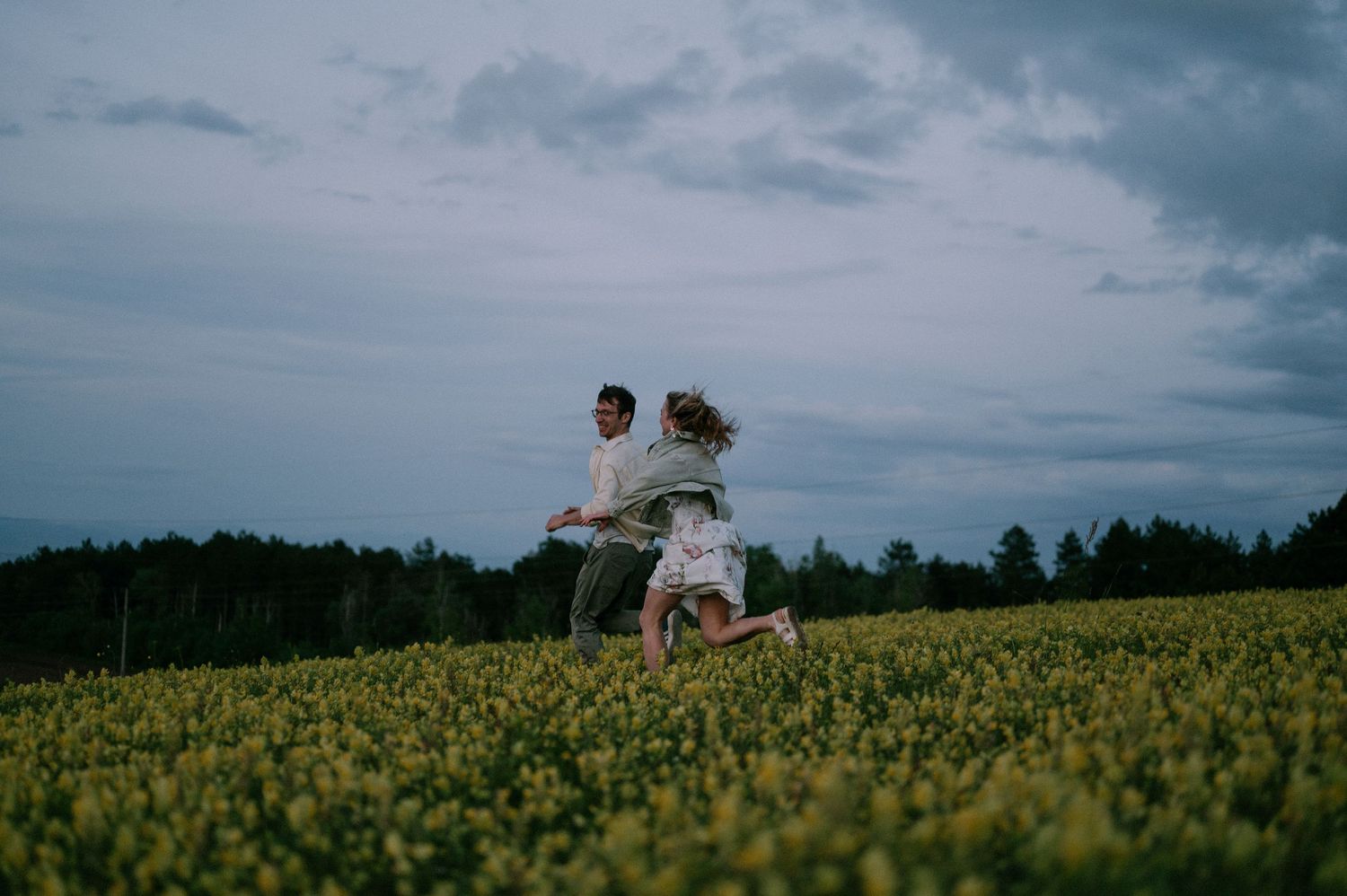 Two figures sit together in a vast yellow canola field under cloudy skies.