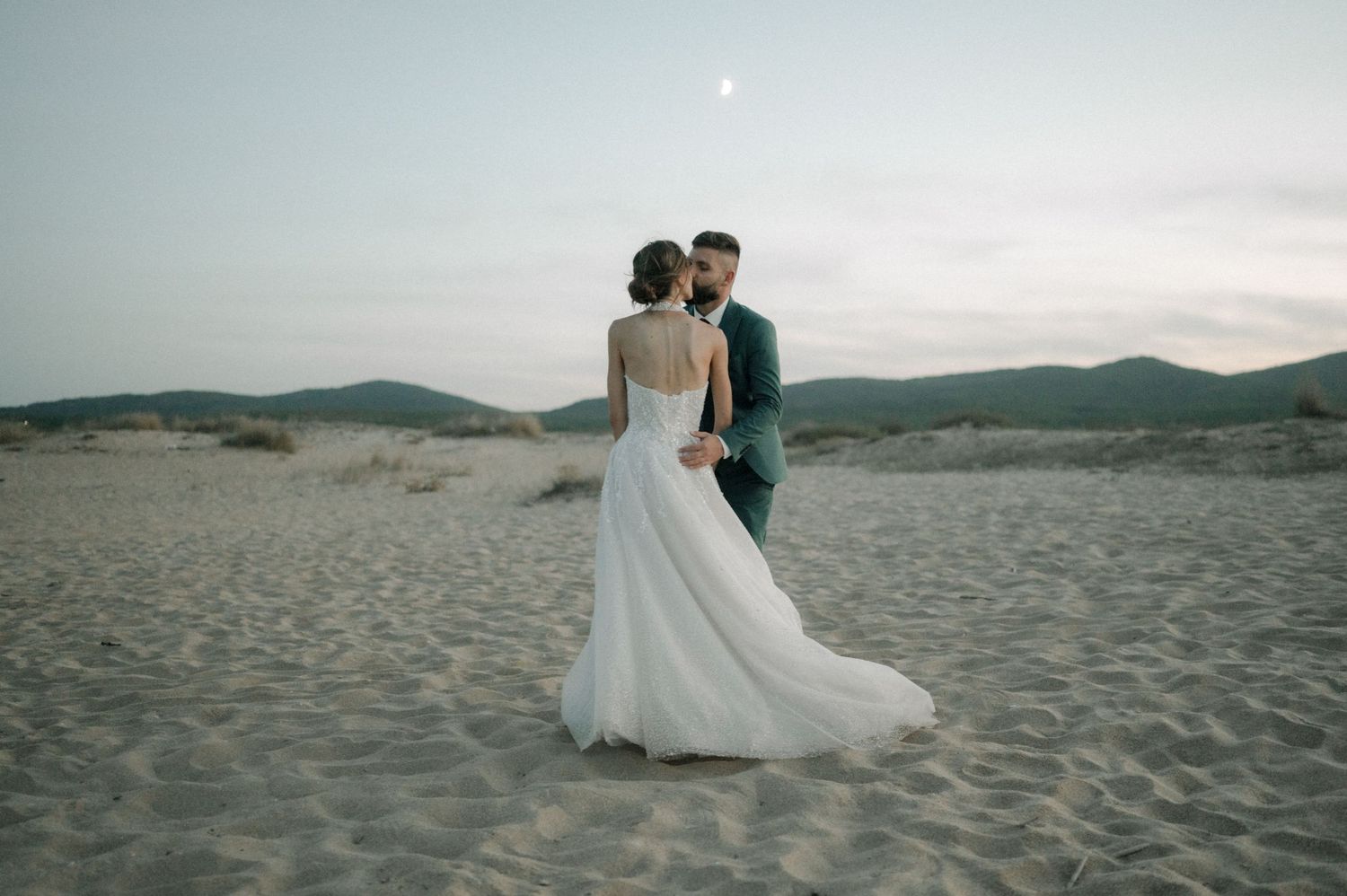 Bride and groom share a romantic moment on sandy dunes at dusk, with mountains and a moon in the background.