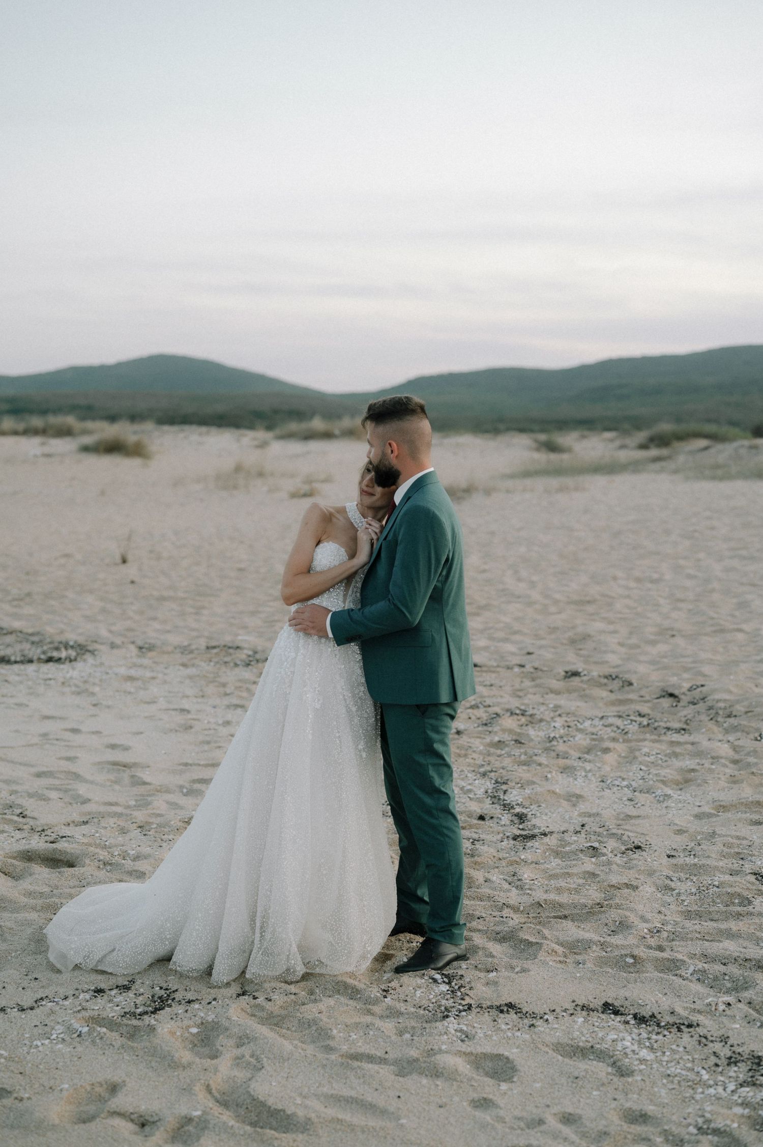 Bride and groom embrace on sandy beach at dusk, she in white gown, he in teal suit, with misty mountains behind.