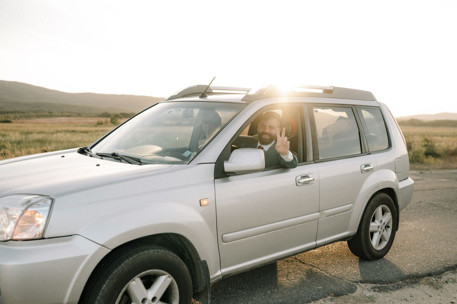 Person waving from silver SUV on scenic road at sunset with golden light illuminating the landscape.