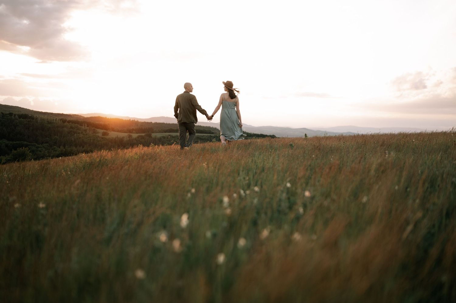 Couple holding hands walking through a lush green meadow at golden sunset with rolling hills in the background.