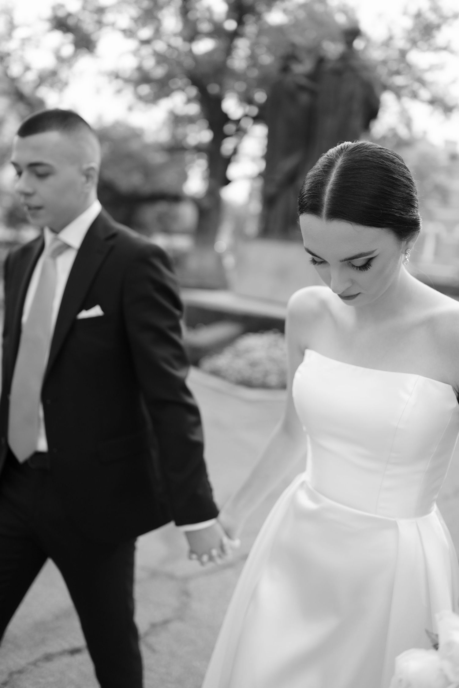 A black and white wedding photo showing a bride in a classic white off-shoulder gown walking with a groom in a dark suit.