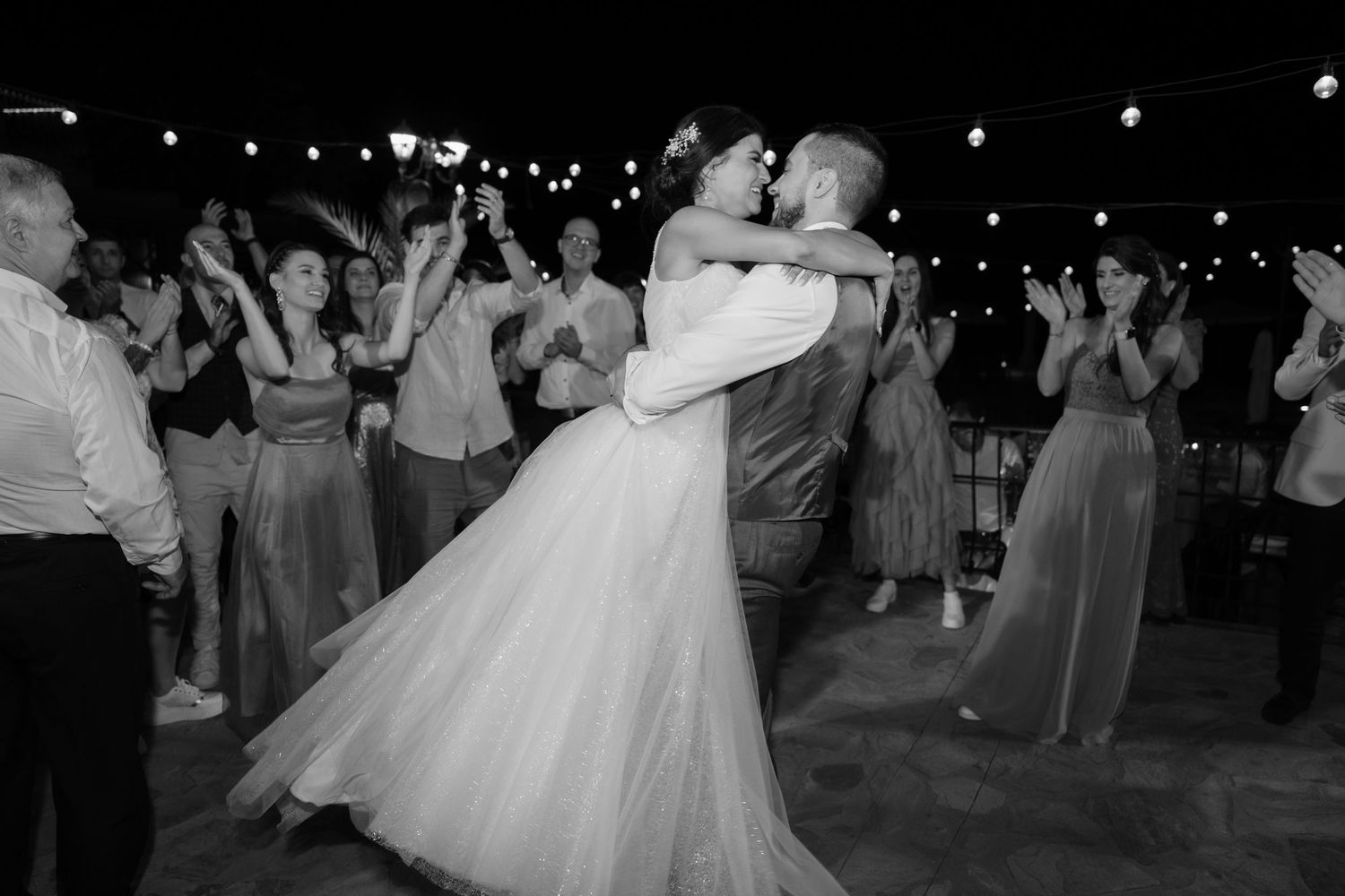 A couple in wedding attire dances together at their reception surrounded by guests and twinkling lights.