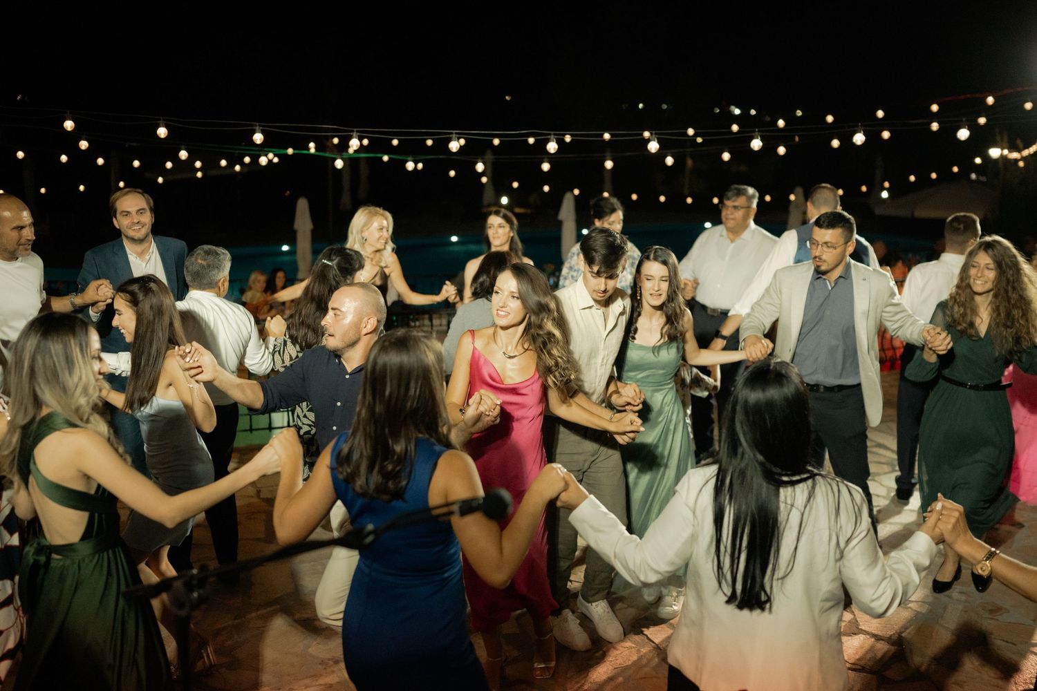 A lively crowd dances together at a nighttime outdoor party with twinkling lights in the background.