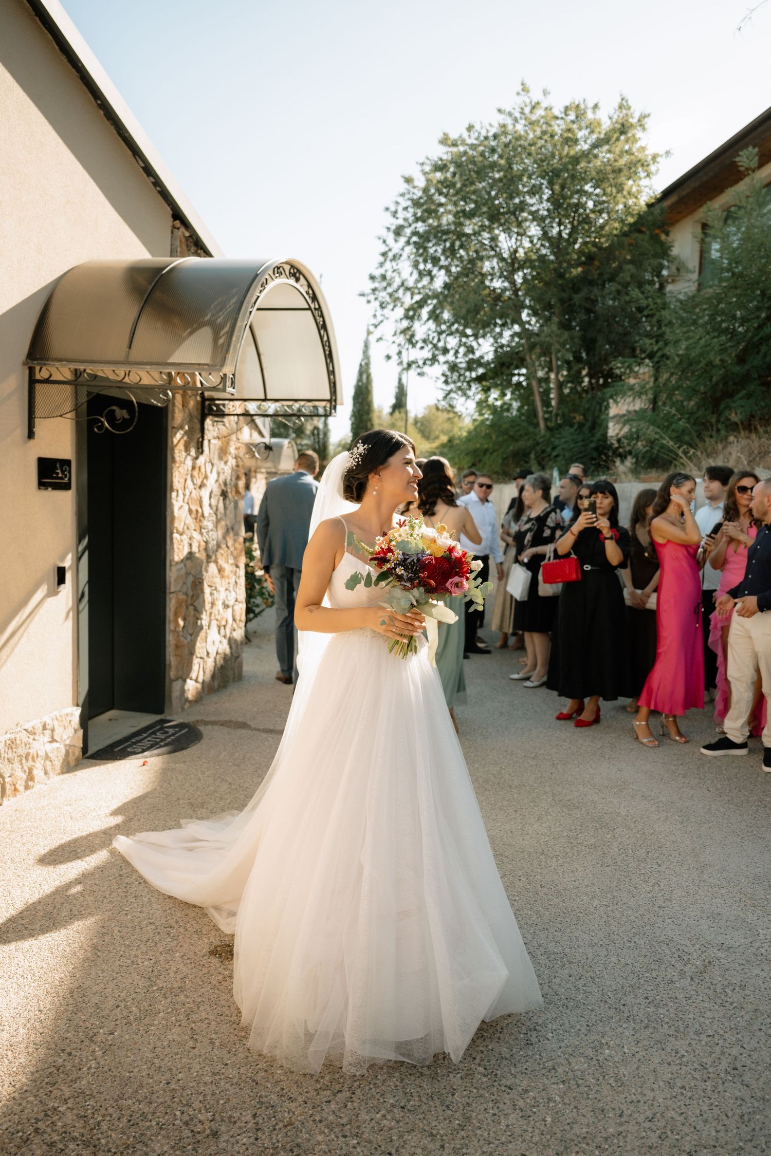A bride in a flowing white gown holds a colorful bouquet outside a stone building while guests look on.