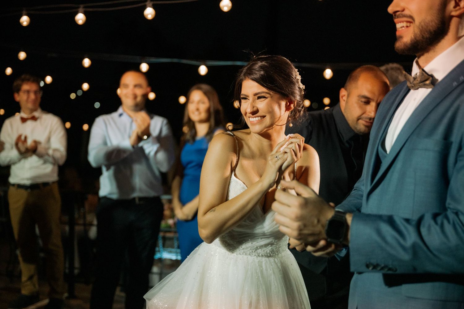 Wedding guests dancing and celebrating at an evening reception with string lights overhead.