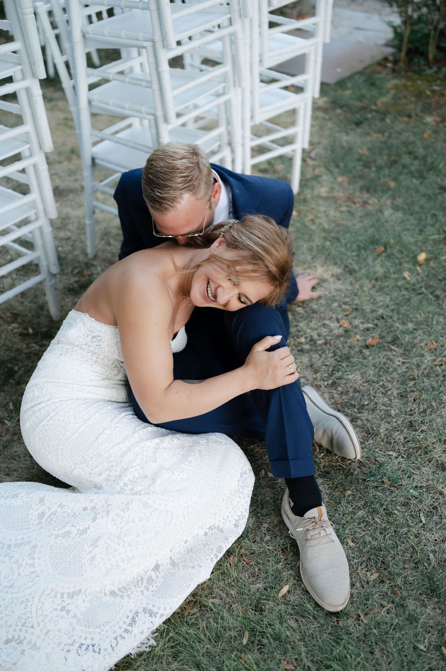 Intimate wedding portrait of couple sharing sweet moments on grass near white chairs.