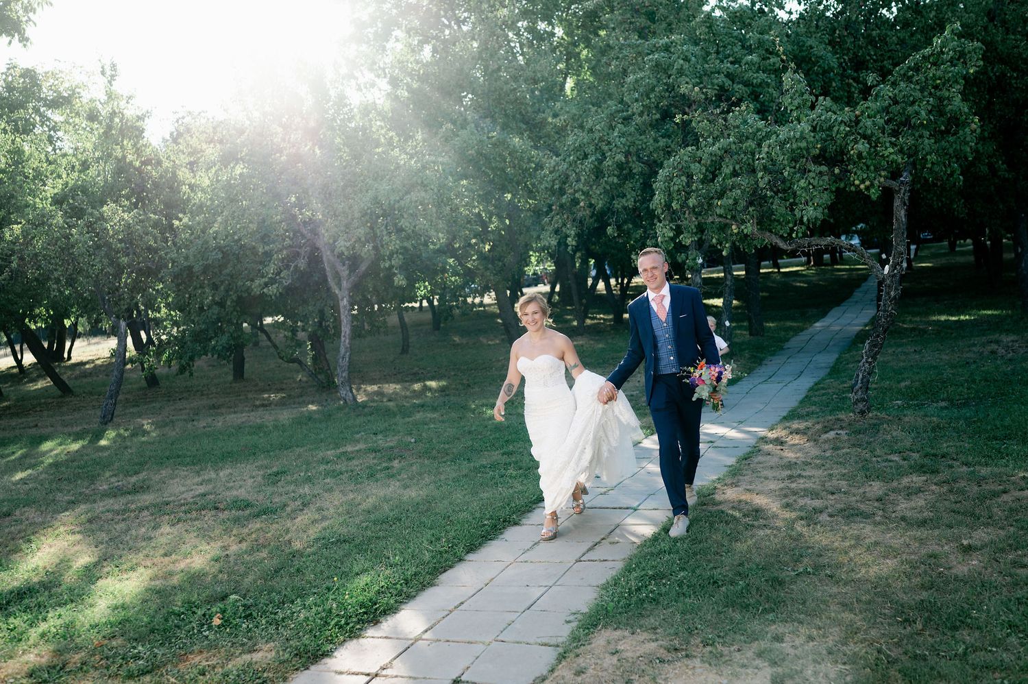 A couple walks along a sunlit garden path surrounded by lush greenery during a wedding photoshoot.