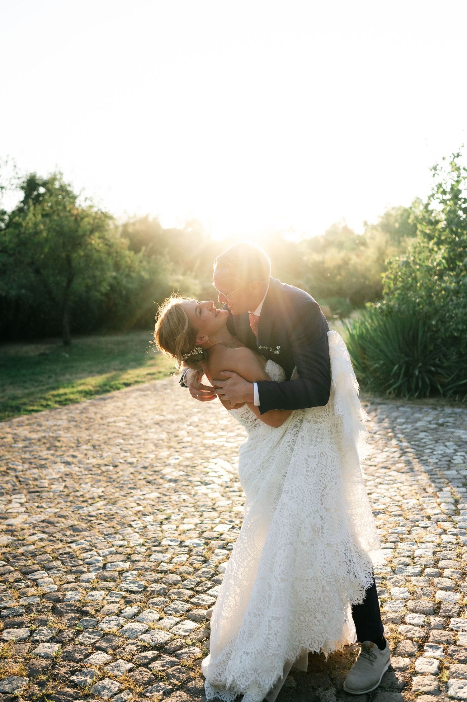 A romantic wedding couple shares a tender moment on a cobblestone path at sunset.