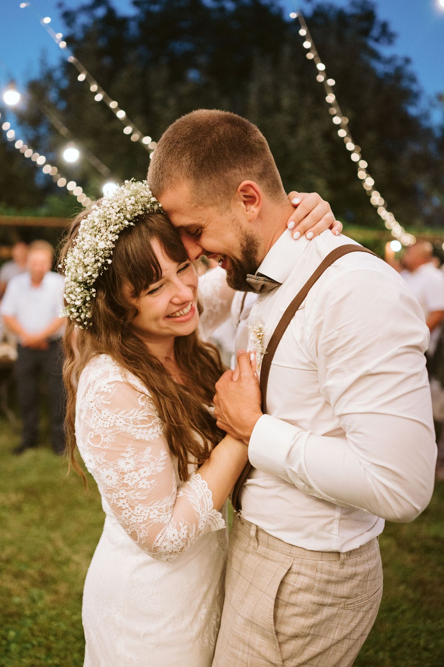 Romantic outdoor wedding dance under twinkling string lights with bride in lace sleeves and groom in light colored suit.