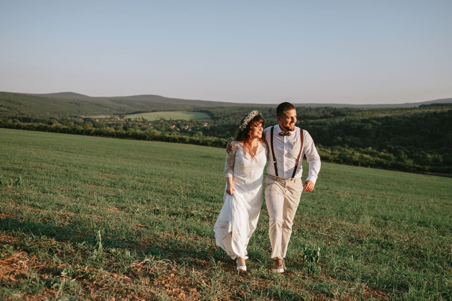 A couple in white wedding clothes walks together through a scenic mountain meadow.