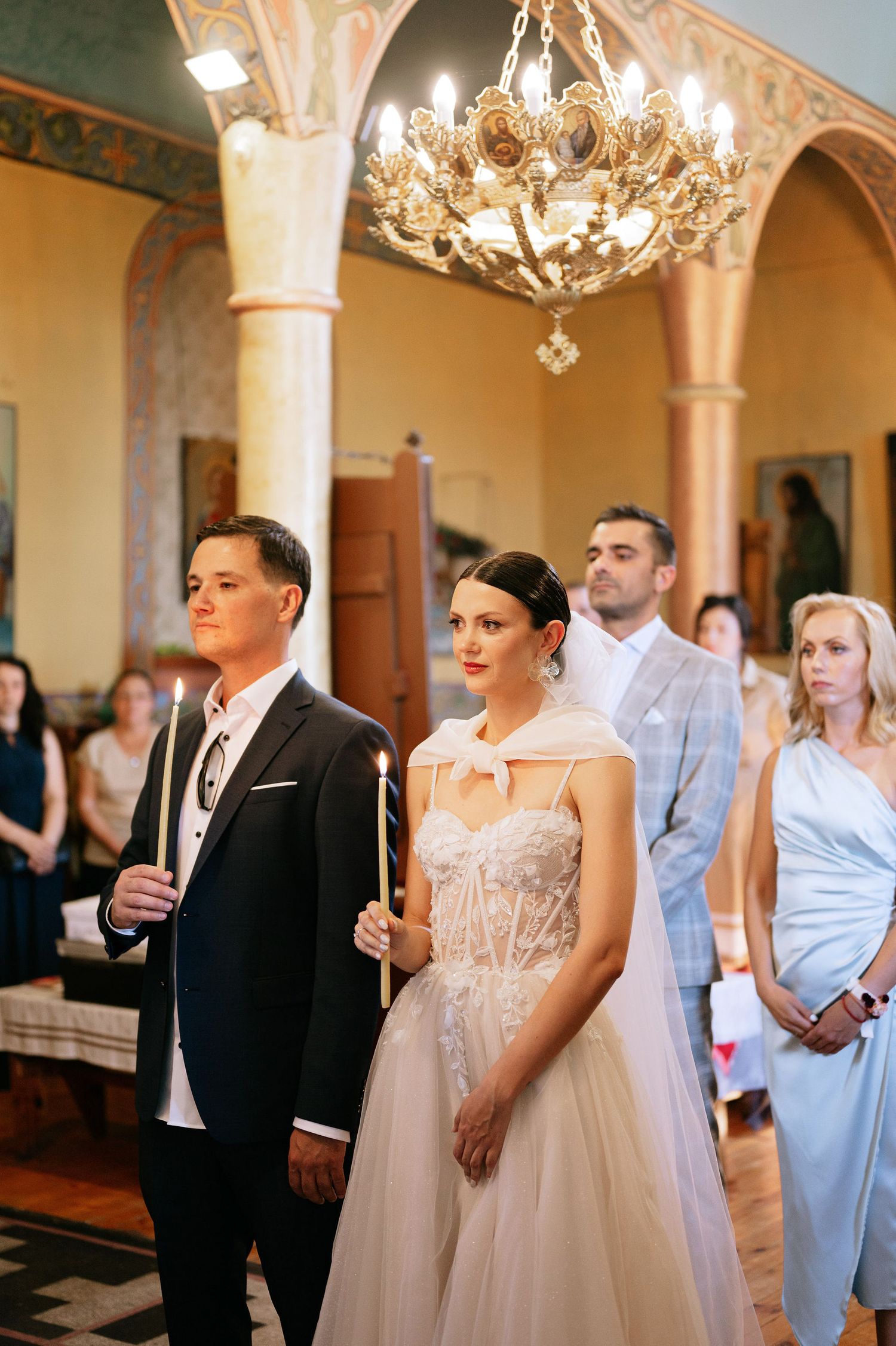 Wedding ceremony participants stand in formal attire inside an ornately decorated church with chandeliers.