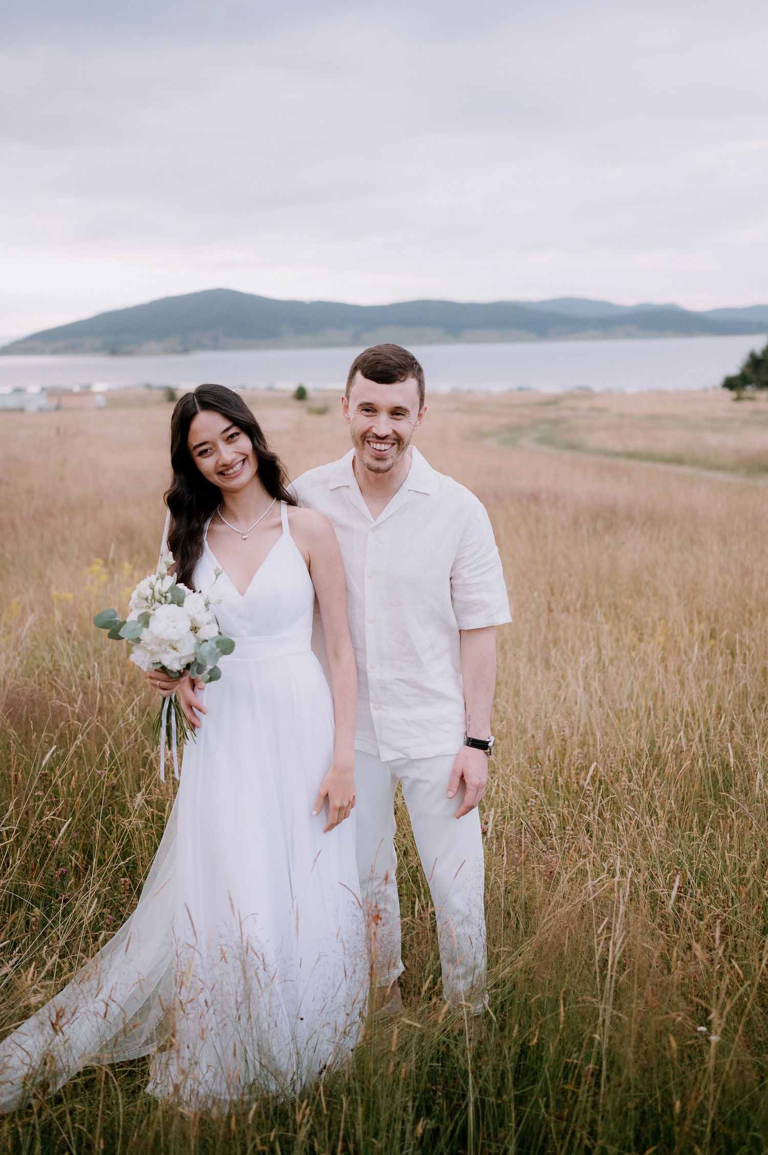A joyful wedding couple poses together in a scenic field with mountains behind them.