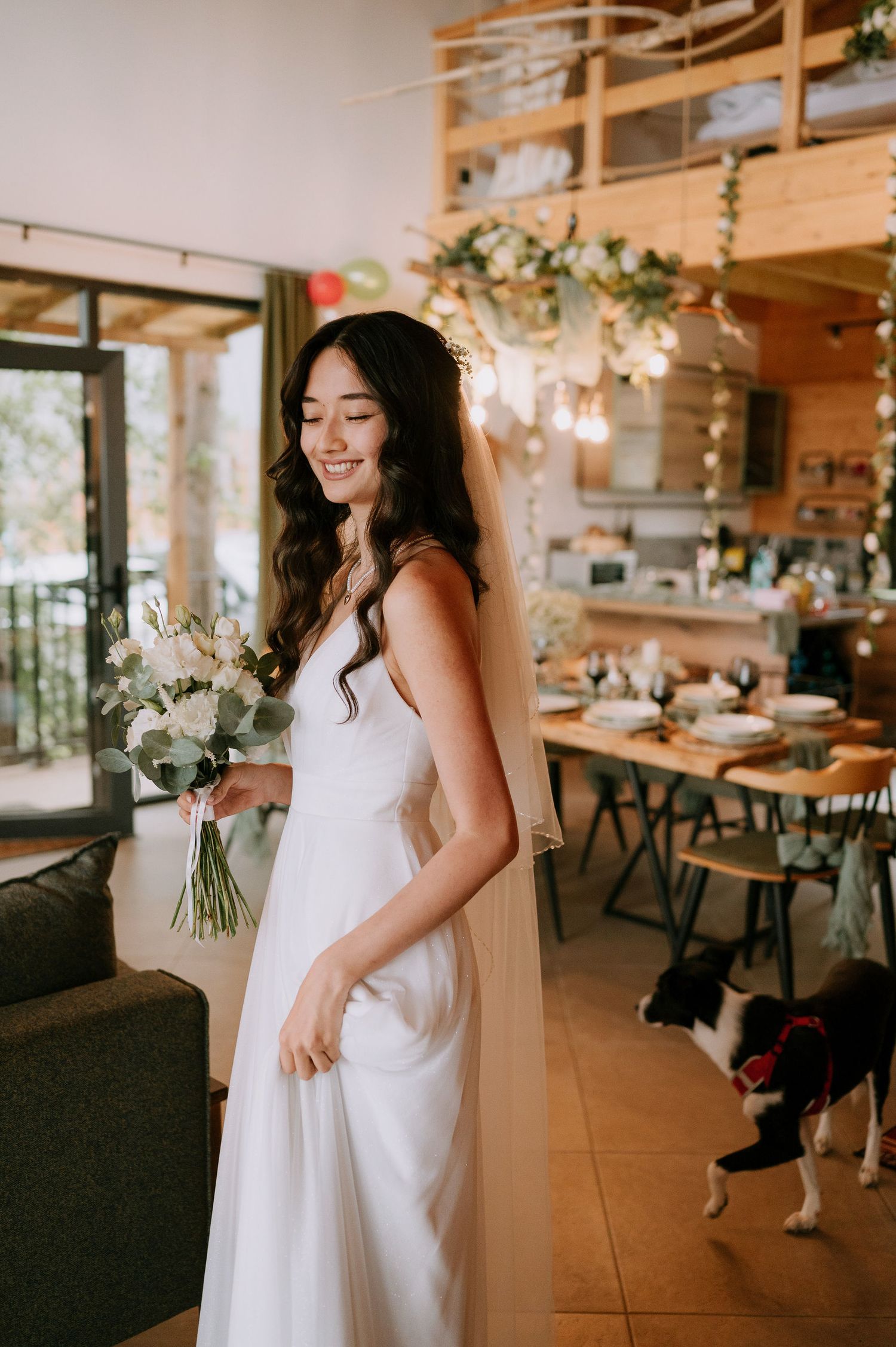 A bride in a white dress holds a bouquet in a rustic wooden venue decorated with string lights.