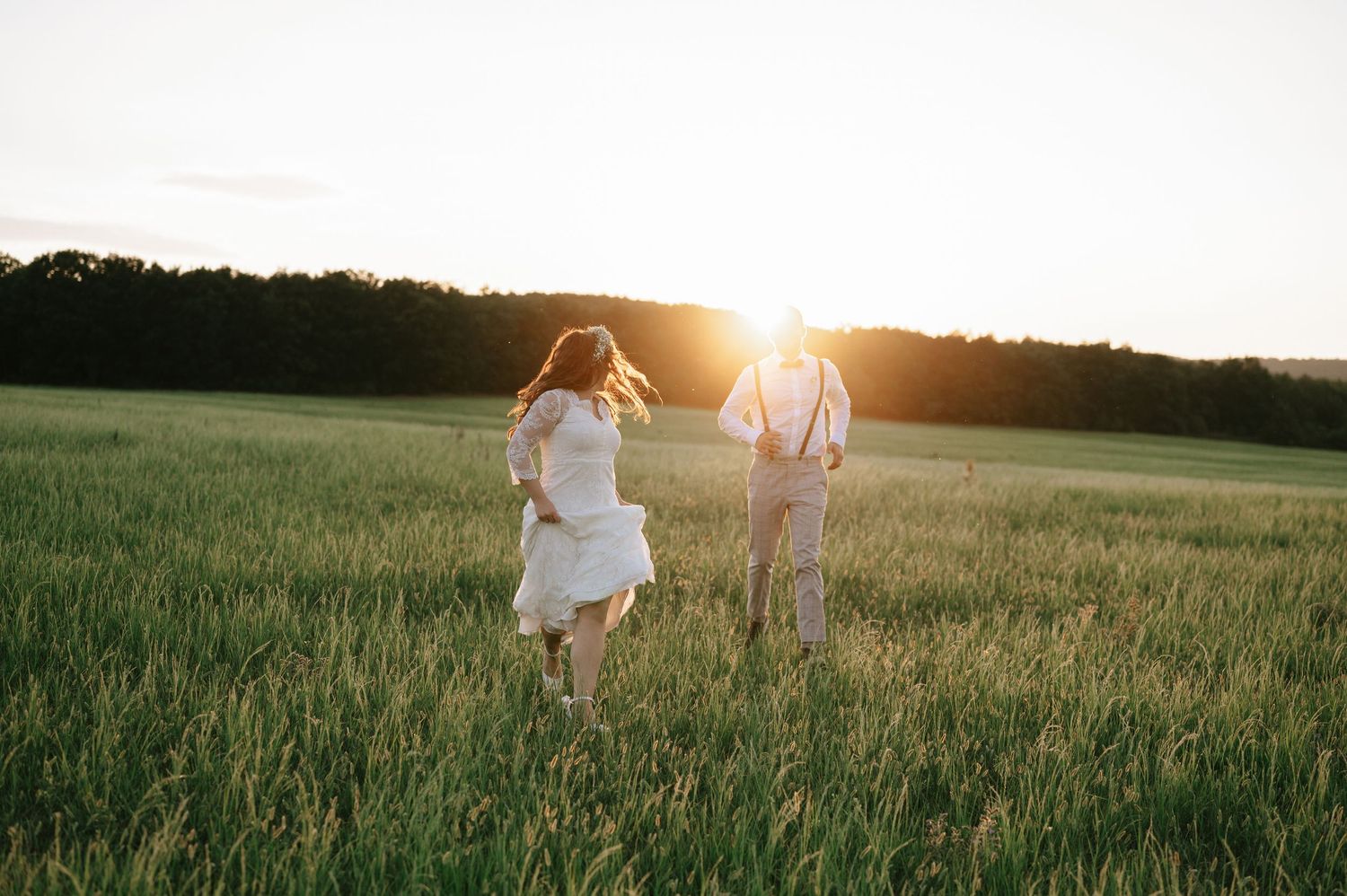 Newlyweds stroll hand in hand through tall grass at sunset.