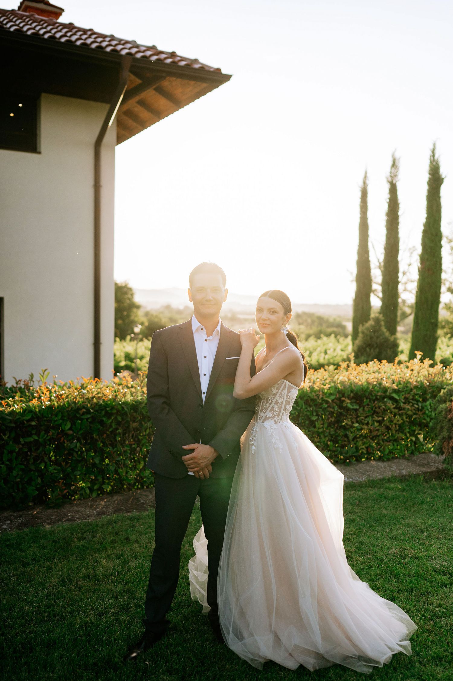 Newlyweds dancing together on lawn during golden hour with white building and cypress trees.