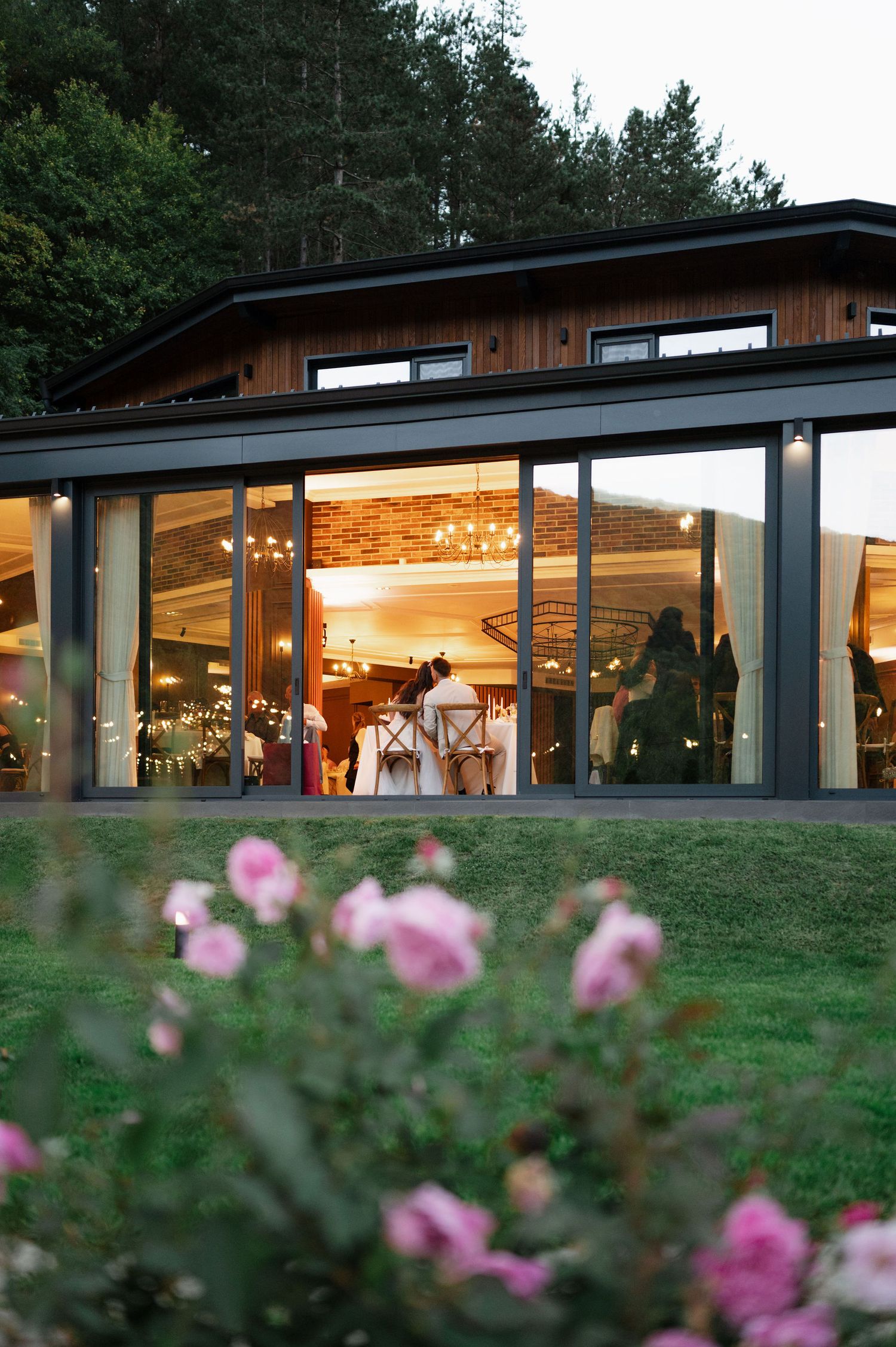 Modern event venue with glass walls illuminated at dusk, framed by pink roses.