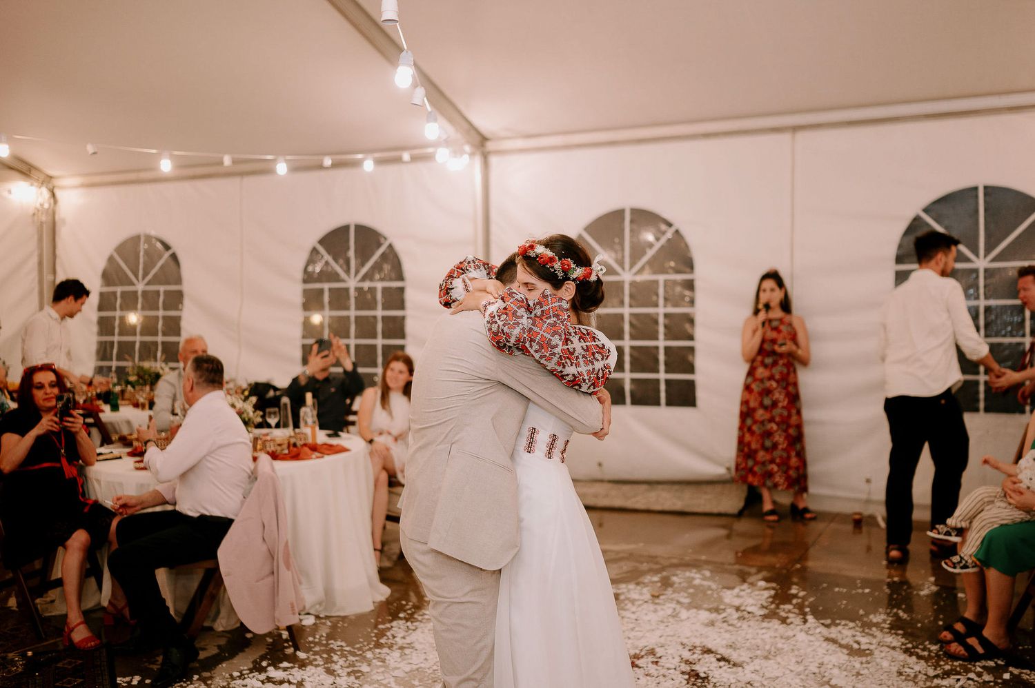 Wedding reception party in full swing under a white tent with decorative lighting and windows.